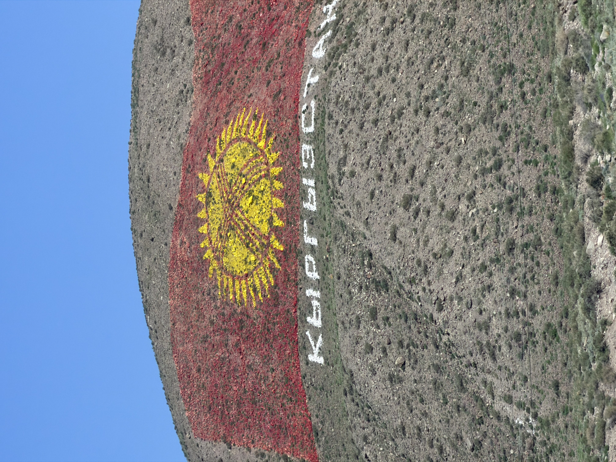 This is an aerial, slightly distorted view of a mountain slope in Kyrgyzstan, featuring the flag of Kyrgyzstan painted onto the rock face. The flag consists of a red background with a yellow sun emblem in the center. To the right of the flag, written in white letters, is the word "КЫРГЫЗСТАН" (Kyrgyzstan) in Cyrillic script. The mountain slope is arid and rocky, with sparse green vegetation. A thin black cable, possibly part of a ski lift or power line, runs diagonally across the mountain face in the upper right portion of the image. The sky is a clear, bright blue. The lighting suggests it is daytime, likely late morning or early afternoon, with no clouds visible. The scene appears peaceful, with the prominent display of national symbols on the natural landscape.