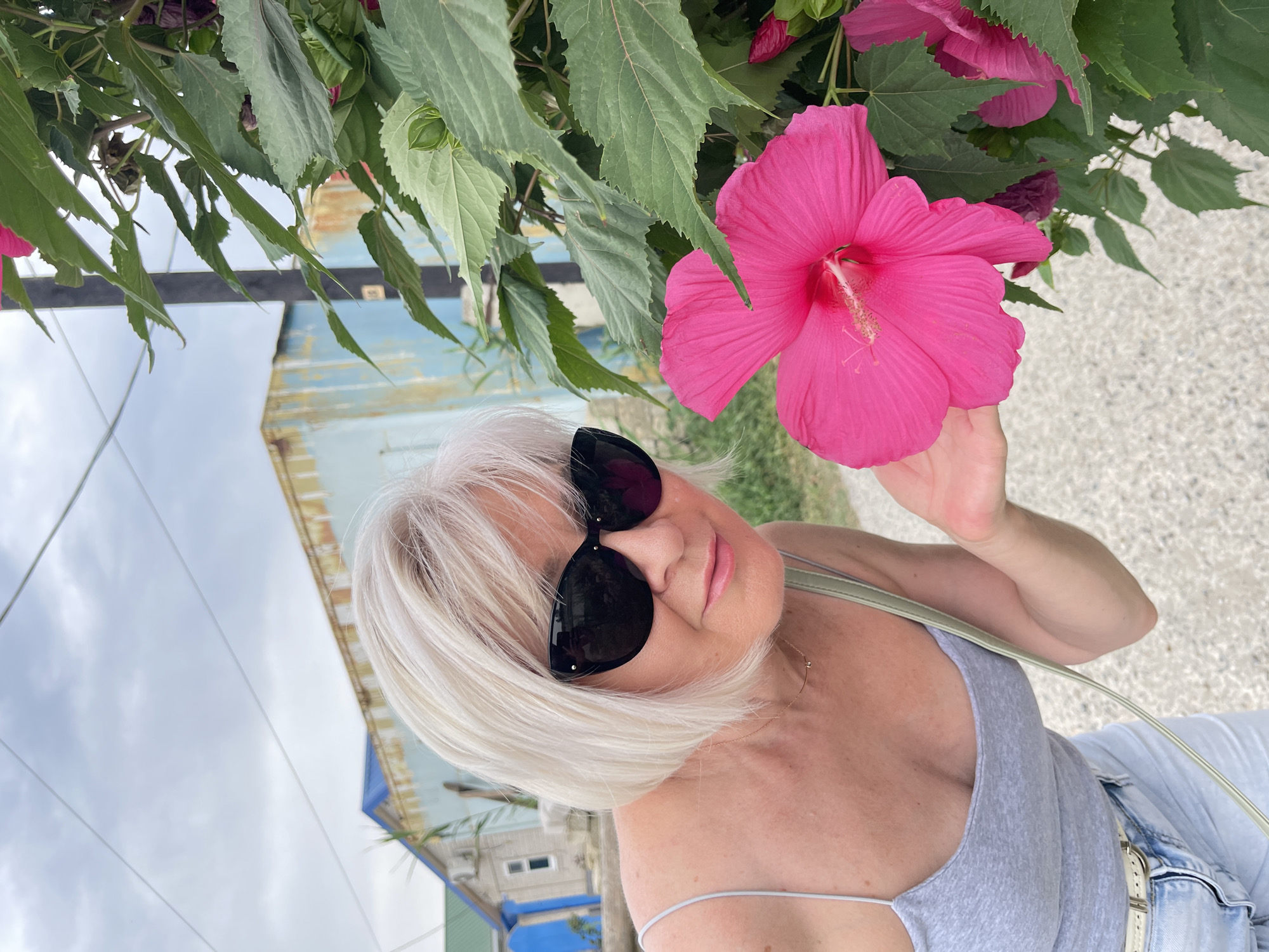 A candid, eye-level, medium shot captures a blonde, middle-aged woman wearing black sunglasses and a gray tank top, leaning in to smell a large, bright pink hibiscus flower. She is positioned on the left side of the frame, with her right hand gently reaching towards the flower. The woman has a light complexion and short, bobbed blonde hair. She is wearing light blue denim shorts, a white belt, and a thin gold necklace. Her left shoulder and part of her chest are visible.
To the right of the woman and dominating the right half of the frame is a hibiscus plant with large green leaves and vibrant pink flowers. The main flower is in sharp focus, its delicate stamen protruding from the center. Several other pink buds and flowers are also visible, some partially obscured by leaves.
In the background, a gray, gravelly path runs horizontally across the lower part of the image. Behind the path and the woman, there are some buildings. The most prominent is a long, light blue building with a weathered texture, possibly a shed or industrial structure. To its left, a smaller, light-colored building with a green roof and a blue door is partially visible. A thin, vertical black pole stands to the right of the woman, partly in front of the blue building. The sky above is overcast with a muted gray cloud cover. The overall setting appears to be outdoors, possibly in a coastal or rural area, consistent with the general description of Pokoinoe, Russia, which is a village located in the Astrakhan Oblast, near the Caspian Sea.
