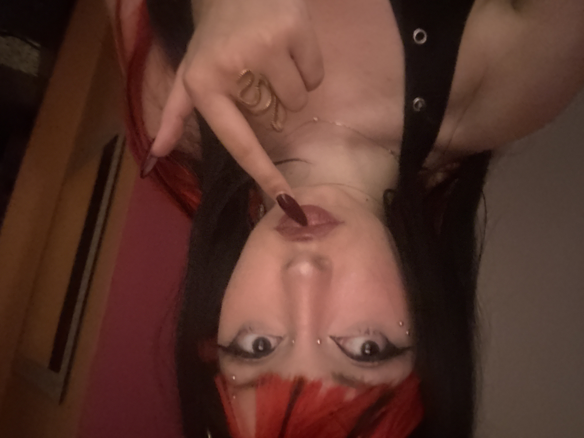 A young woman with red-streaked black hair, facial piercings above each eyebrow, and dark eye makeup poses for a selfie. She wears a black tank top with silver grommets and a gold snake ring on her hand. Her finger touches her lips. The background is out of focus, but suggests an indoor setting, possibly a room with a pink wall and a wooden frame. The image does not show any characteristics to indicate the location is in Milan, Italy.