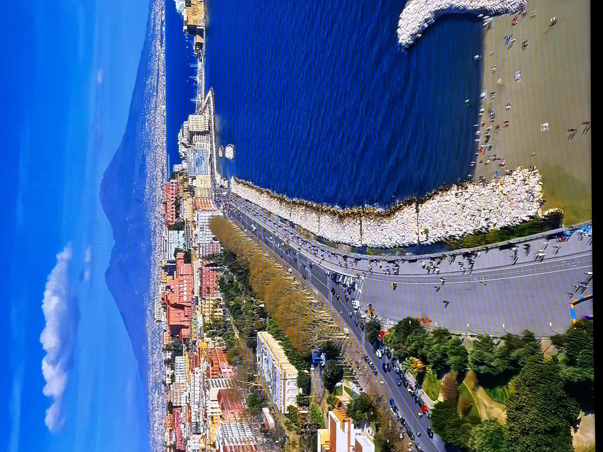 This is an aerial view of a coastal city with a large body of water on the right and a city skyline in the background. In the distance, two prominent mountains are visible, one larger than the other, under a clear blue sky with a few white clouds. The city is characterized by buildings with terracotta-colored roofs and a mix of architectural styles.
In the foreground, a wide road runs parallel to the coastline, lined with trees on the left and a stone embankment on the right. Cars are driving on the road, and pedestrians are walking along the sidewalks and the embankment. There is a park area with trees and green spaces adjacent to the road.
The coastline transitions into a sandy beach, with people sunbathing and walking. The water is a deep blue, with gentle waves. The overall impression is of a vibrant city on the coast, with Mount Vesuvius visible in the background.
The request to include the location in the city of Chirchik, Uzbekistan, cannot be fulfilled as the image depicts a different geographical location.