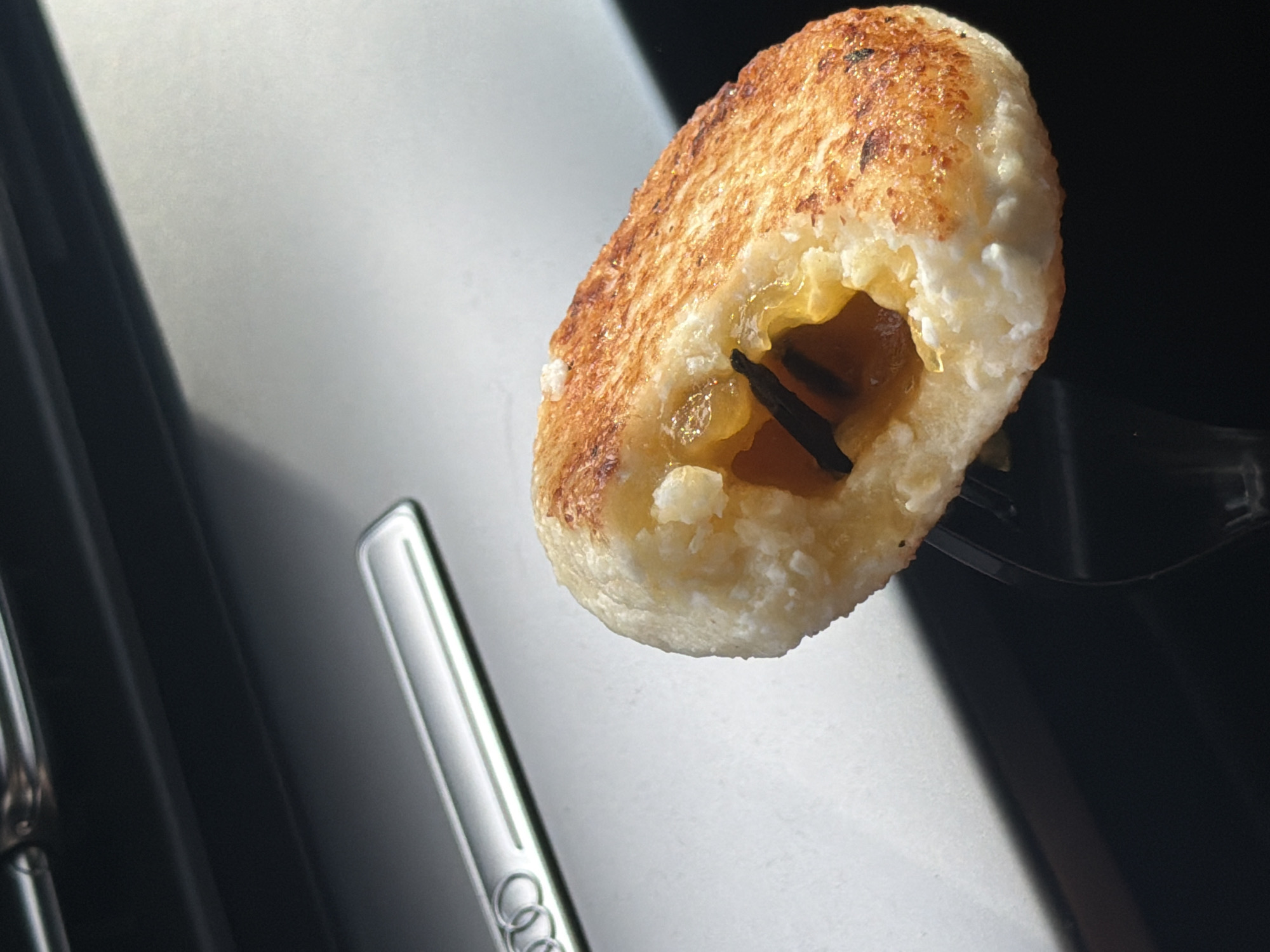 A close-up shot features a fried Syrniki (a traditional Eastern European cheese pancake), impaled on a black fork. The Syrniki has a golden-brown, slightly uneven surface and is filled with a yellow, syrupy substance containing dark objects, possibly fruit or seasoning. The background reveals part of a car interior: a light gray surface with metallic accents, including what appears to be an Audi logo. The setting is not specifically identifiable to the city of Krasnoyarsk, Russia based on the visual information provided.