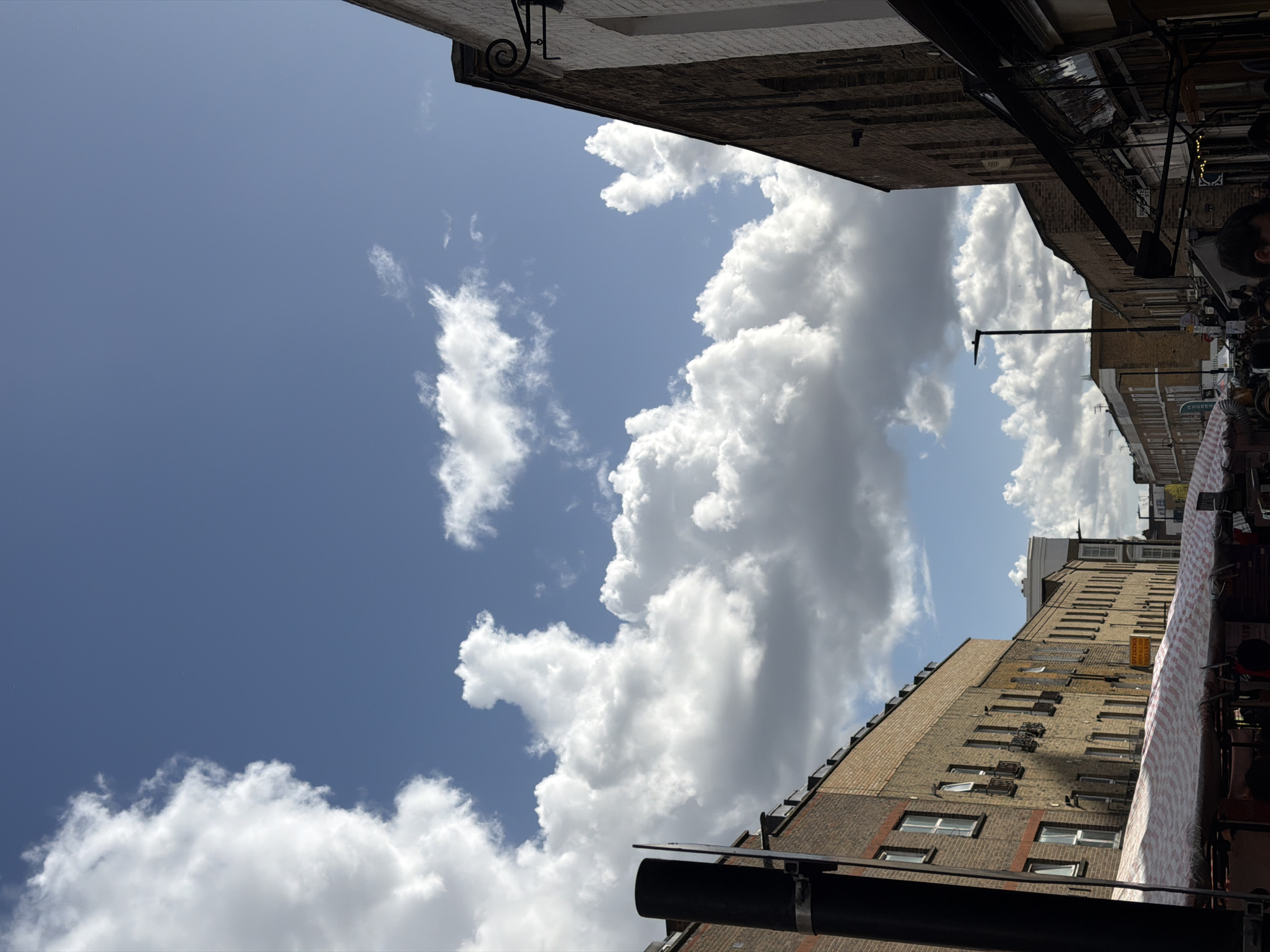 The image depicts a view upwards, looking at buildings and a partly cloudy sky in London, UK. Brick buildings frame the shot on either side, with windows and various architectural details visible. Overhead, awnings and hanging objects suggest a street market or commercial area. Bright white cumulus clouds fill a portion of the blue sky. Signs and a lamppost are discernible among the structures. The lighting suggests a sunny day.