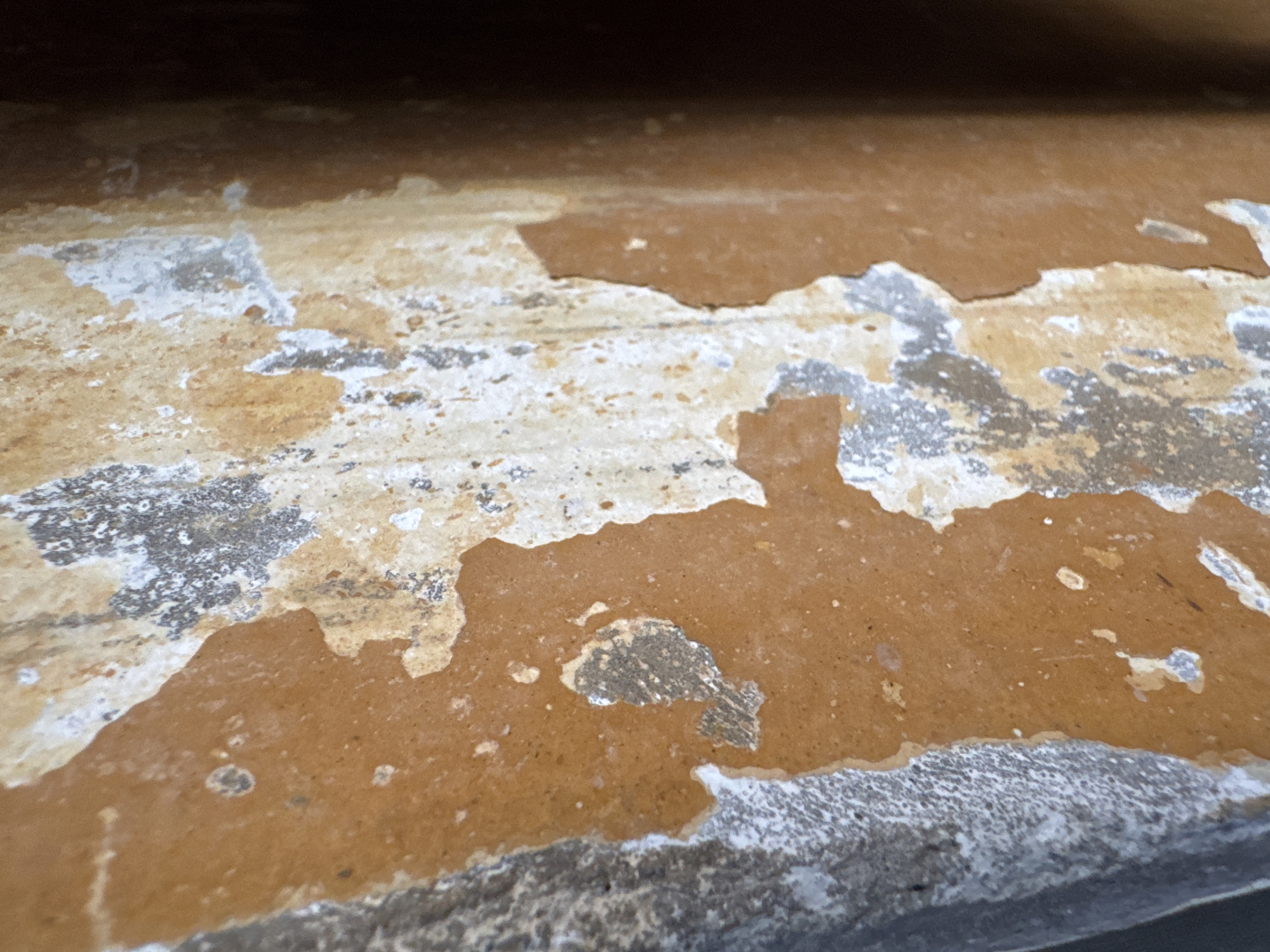 The image shows a close-up of a surface with peeling paint. The top layer is a brownish-orange color, and beneath it, there are patches of white and gray. The surface appears to be rough and textured, with the peeling paint creating an uneven appearance. The image is taken from a low angle, giving a perspective that emphasizes the textures and colors of the peeling paint. The background is dark and out of focus, which helps to isolate the peeling paint as the main subject of the image. There are no people or other objects visible in the image.