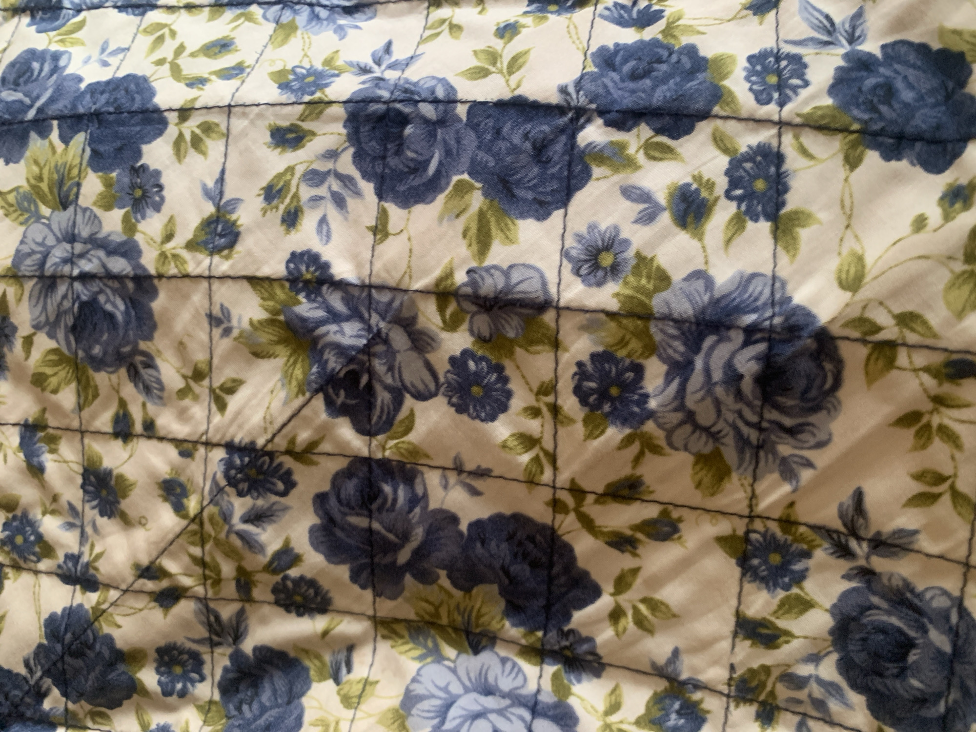 The image displays a close-up, overhead view of a quilted fabric with a blue floral pattern. The fabric has a cream-colored background with various shades of blue roses and smaller blue flowers, interspersed with green leaves. The quilted pattern creates a grid-like texture across the fabric, with visible stitching lines forming squares. The lighting appears to be natural, casting subtle shadows and highlighting the texture of the quilt. The scene is devoid of any people or specific environmental context that would allow for precise location identification within Jalingo, Nigeria. The focus is solely on the pattern and texture of the textile.