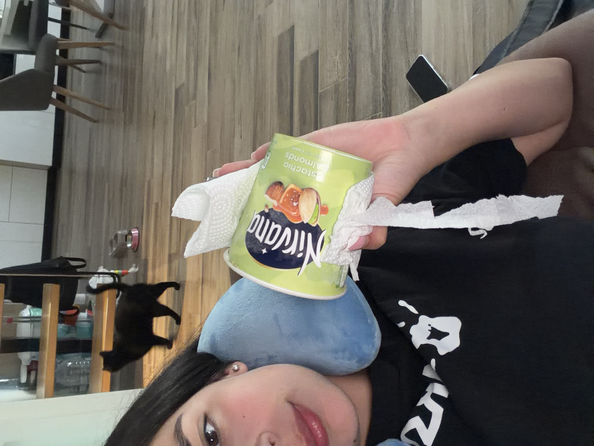 A woman is holding a container of Nirvana Pistachio & Almonds ice cream with a paper towel wrapped around it. She has a blue travel pillow under her head. The woman is wearing a black shirt with white designs. A black cat is standing on a white shelf. A smartphone is visible in the background on a wooden floor. Based on the brand of ice cream, this image is likely not in Gabrovo, Bulgaria.