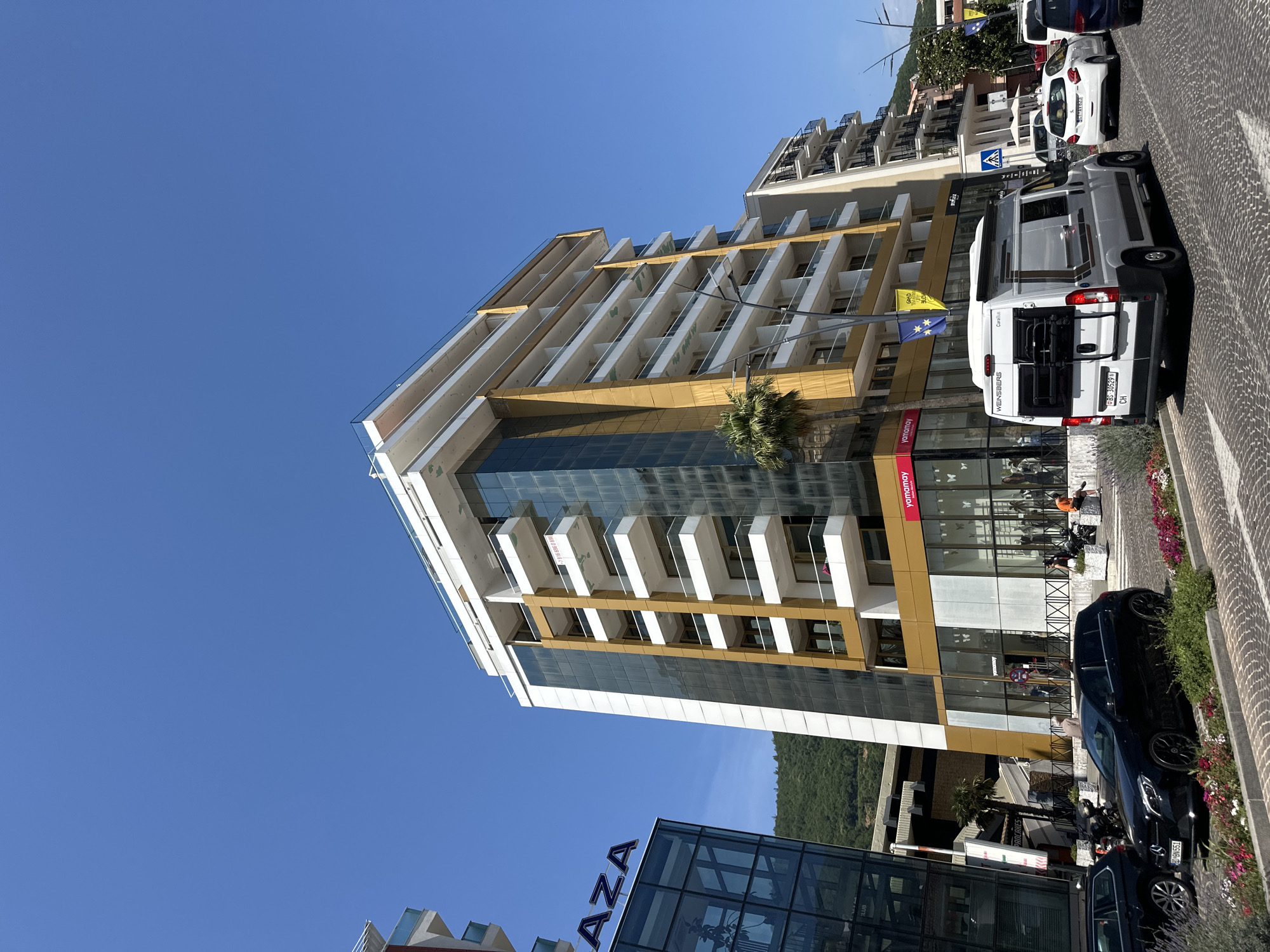 The image depicts a street scene in Budva, Montenegro, likely near the coast, on a clear, sunny day. A modern, multi-story building with a golden facade and white balconies dominates the foreground. To the left, a glass-fronted building with the word "PLAZA" is visible. Several cars are parked on the cobblestone street, including a black Mercedes hatchback and a white campervan. Some people are visible on the sidewalk and entering the building. The overall impression is of a developed, urban environment with contemporary architecture.