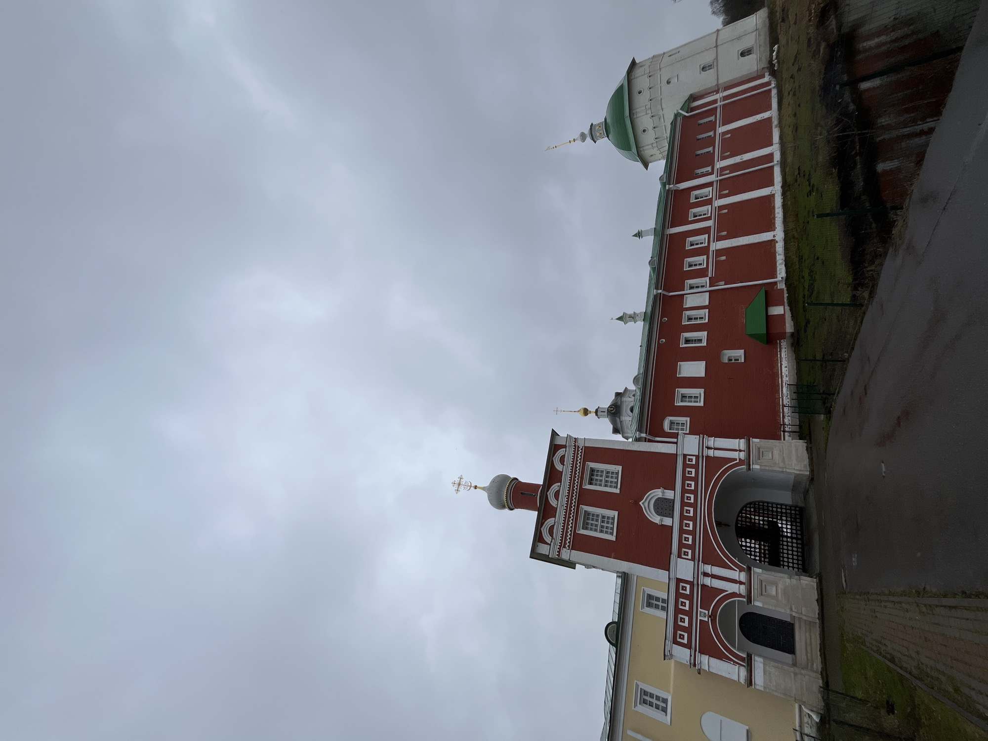 The image depicts a complex of buildings in Луговой, Russia under an overcast sky. The buildings are predominantly red brick with white trim and feature various architectural elements, including a tower topped with a green dome and spire, a gateway with a black iron gate, and other towers with domes and crosses. A tall, white building with a dome is visible in the background, possibly part of the same complex. There is a patch of green vegetation on the slope next to the red brick building. A paved road or walkway runs along the edge of the frame.