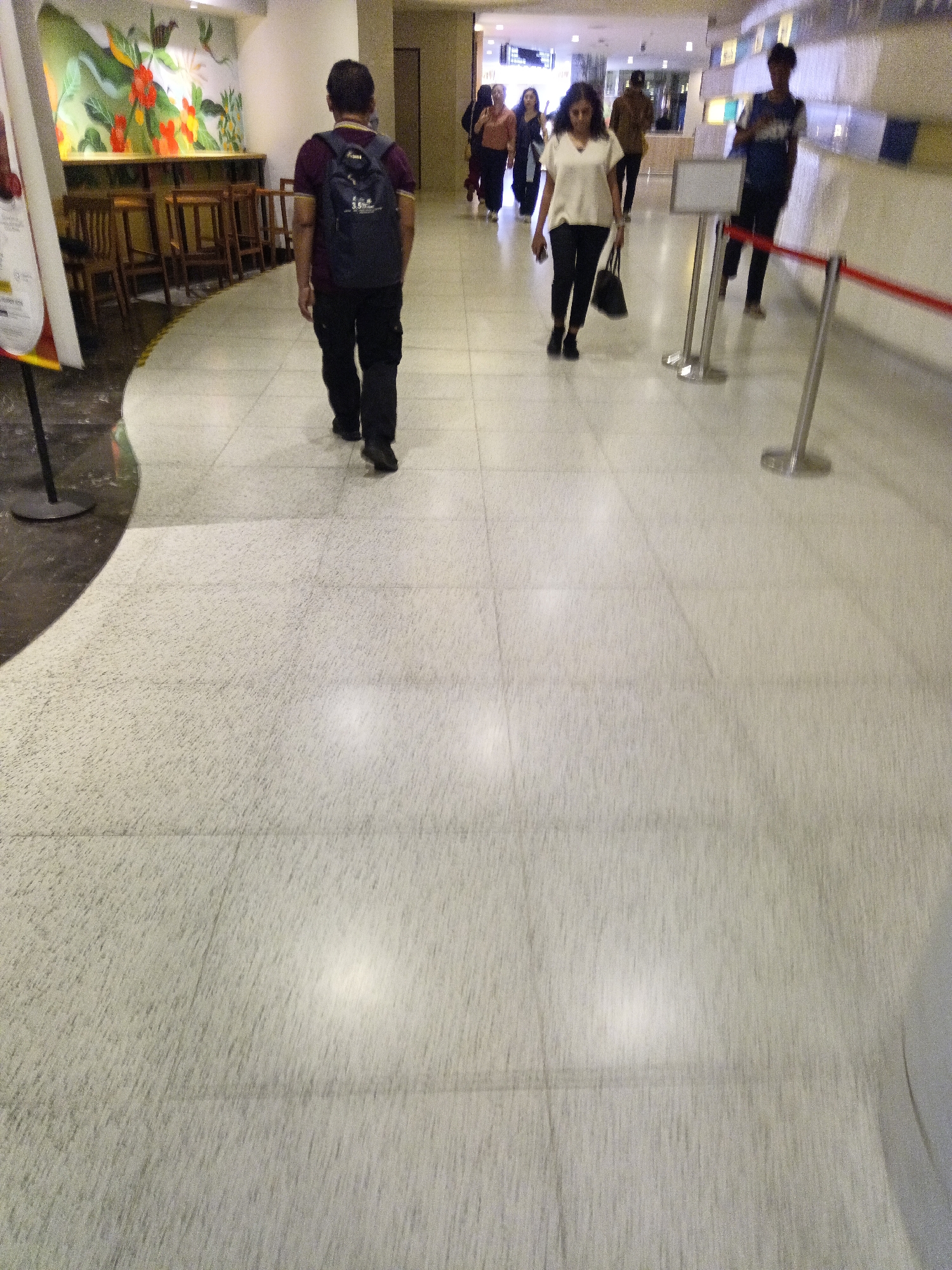 The image depicts an indoor walkway with a light-colored, speckled tile floor. In the foreground and midground, several individuals are walking away from the camera. A man in a maroon shirt and dark pants with a backpack is walking in the left midground. Further ahead, a woman in a white top and black pants with a black handbag is walking. Behind her, a man in a brown shirt and dark pants is also walking. In the background, other people are visible in a blurred state, and there's a counter area with signage. The environment suggests a public indoor space, possibly a shopping mall or an airport in Kuala Lumpur, Malaysia, given the presence of people of diverse ethnicities common in the region. The lighting appears to be artificial, casting a slight glare on the floor.