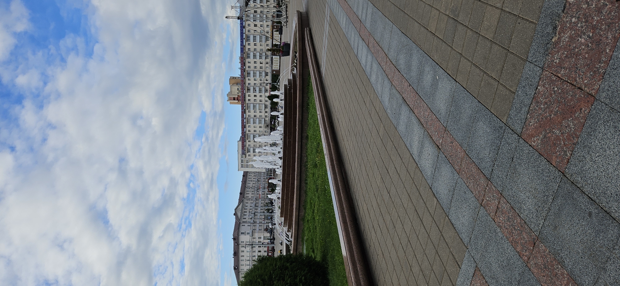 The image depicts Victory Square in Vitsebsk, Belarus, on a cloudy day. In the foreground, a paved walkway with alternating granite and stone tiles leads the eye towards the center of the square. A circular fountain with multiple jets of water is the focal point of the mid-ground, surrounded by a manicured lawn and a low granite retaining wall. Behind the fountain, several multi-story buildings line the square, their architecture suggesting a Soviet-era influence. One building prominently displays a large sign with text in Cyrillic, reading "ПЕРВОЕ ПРАВИЛО ПРИ ЧРЕЗВЫЧАЙНОЙ СИТУАЦИИ - НЕ ПАНИКОВАТЬ" (The first rule in an emergency is not to panic). The sky is filled with a mix of white and grey clouds, with patches of blue sky visible. There are no people or vehicles clearly visible in the frame. The overall scene conveys a sense of urban tranquility and civic space.