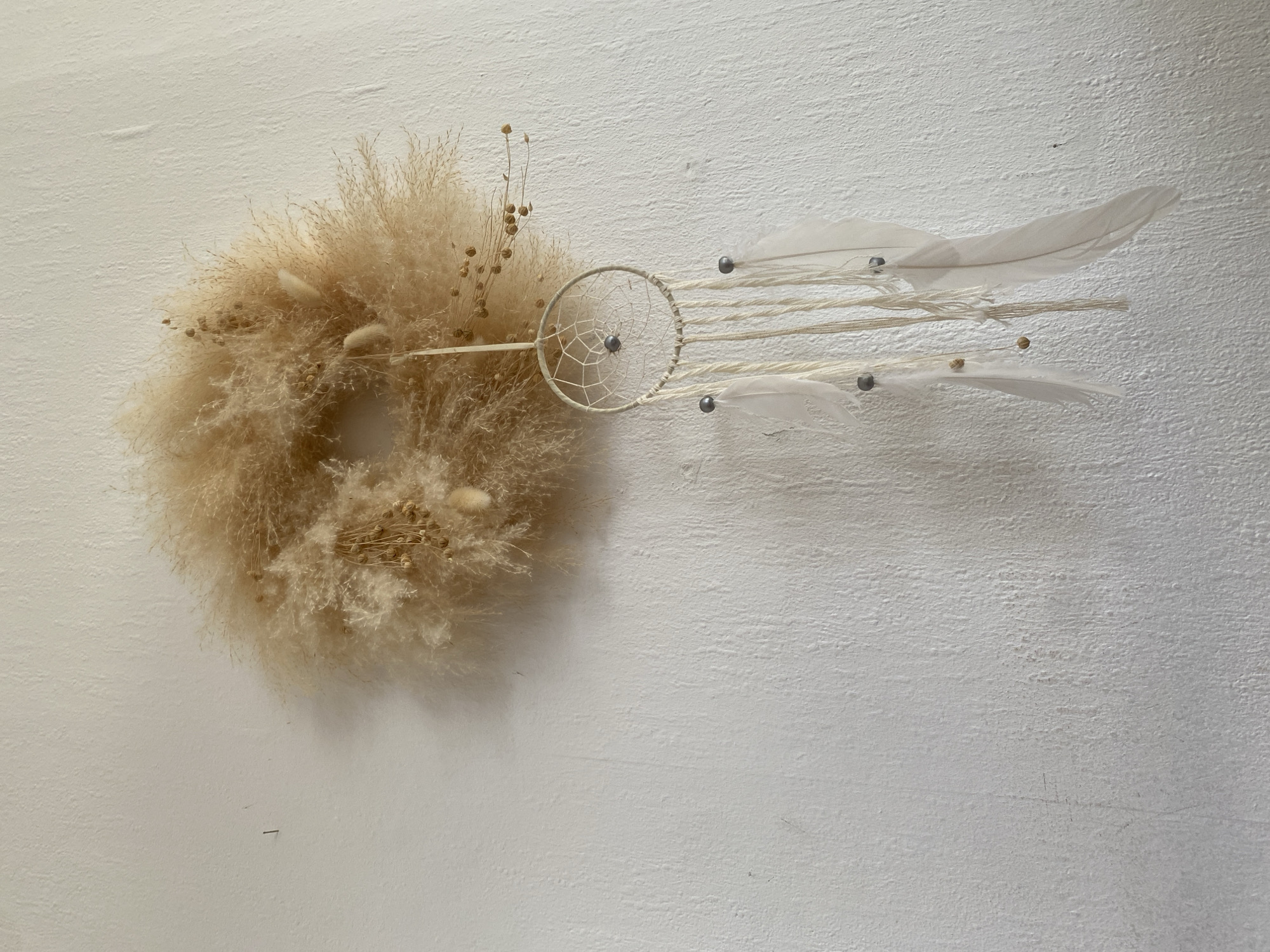 A decorative wreath made of dried grass and small seed pods hangs on a textured white wall. Attached to the bottom of the wreath by a thin string is a dreamcatcher. The dreamcatcher has a circular frame woven with white string, a central bead, and several white strings hanging down, adorned with beads and white feathers. The scene is captured from a medium distance, with the objects occupying the upper right portion of the frame. The lighting suggests an indoor setting, possibly a home, with a slight shadow cast by the wreath and dreamcatcher on the wall. The environment appears to be a domestic interior in Bad Sachsa, Germany.