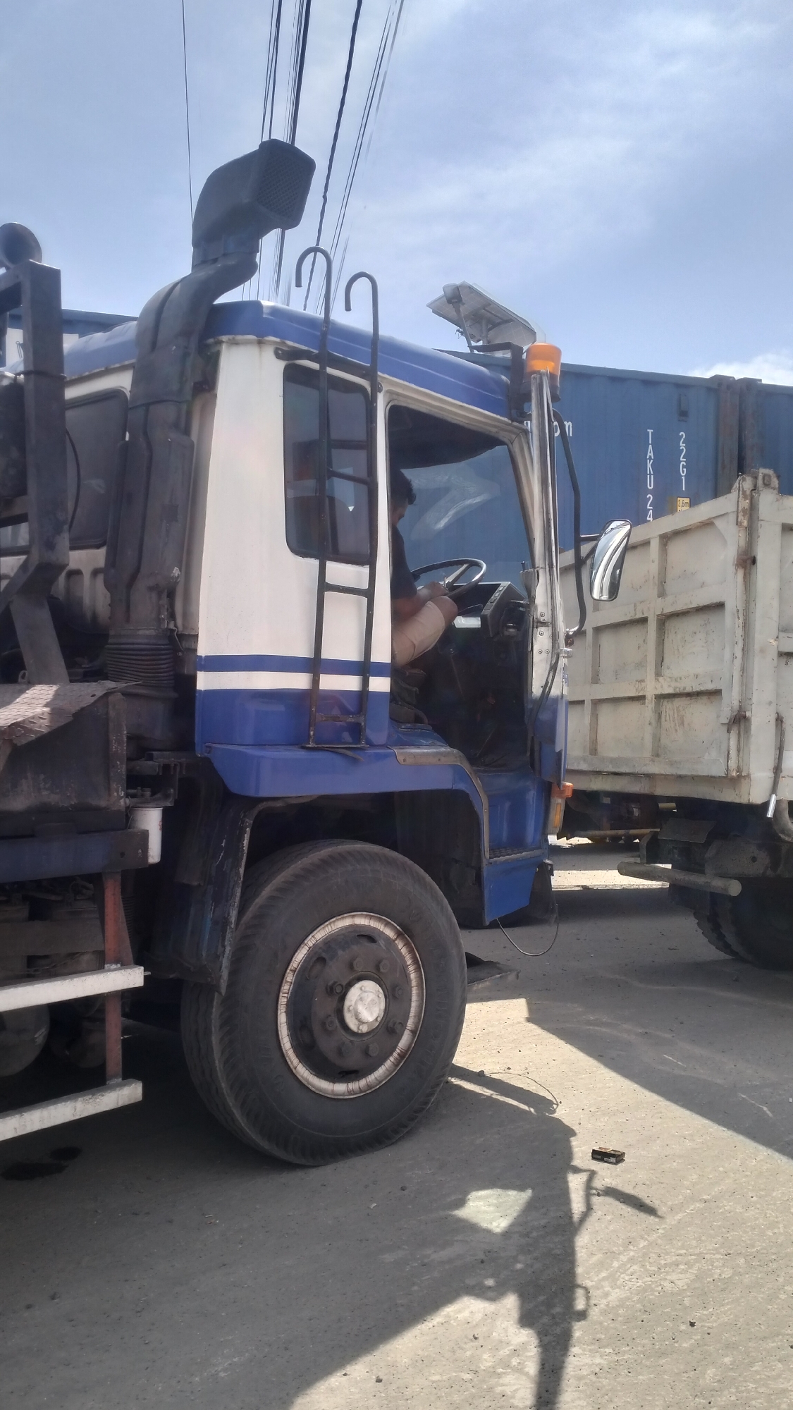 The image shows a large blue and white truck parked on a street in Lhokseumawe, Indonesia. The truck has a snorkel air intake on the left side and a ladder leading to the cabin door, which is open. A person is visible inside the cabin, seated behind the steering wheel. The truck is partially obscured by a white trailer attached to its rear. In the background, a blue shipping container is visible, with some text and numbers on its side, including "TAKU 24" and "22G1". The sky is partly cloudy, and the ground is paved. The overall scene suggests a typical urban street environment in Indonesia.
