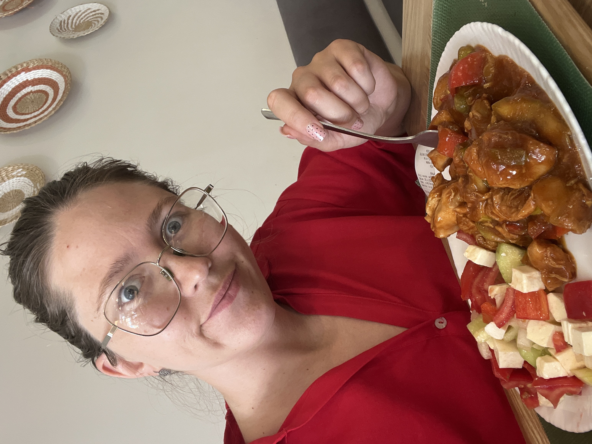 A woman with glasses and a red shirt is holding a fork in her right hand and posing for a selfie next to a plate of food, which consists of meat with a sweet and sour sauce, a salad with tomatoes, cucumbers, and white cheese. The photo was taken indoors, near a wall decorated with woven baskets. The plate is resting on a green and brown surface, possibly a placemat or tray. Based on the presence of the receipt, the photo was taken at the restaurant "Racic" in Podgorica, Montenegro.