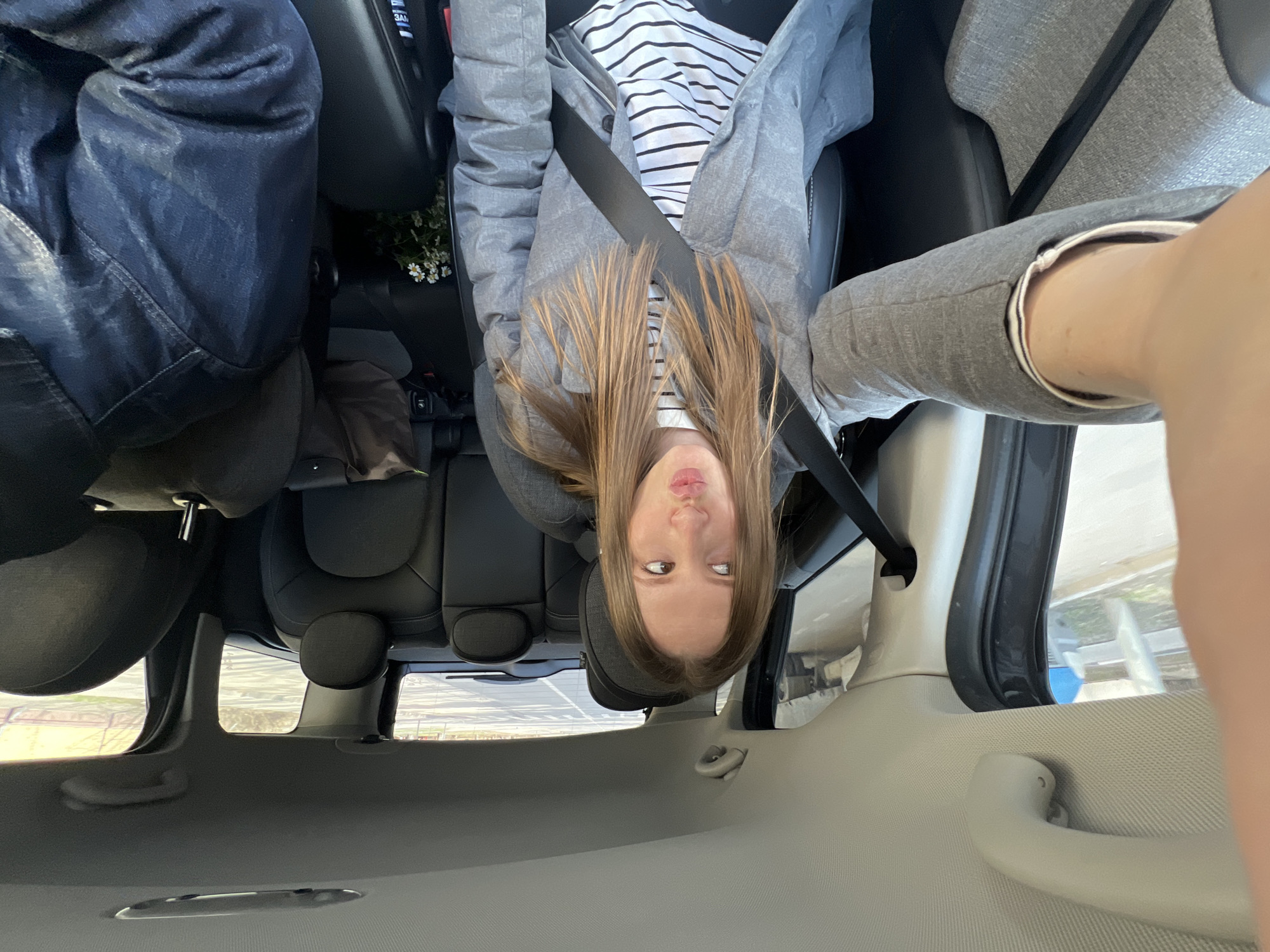 A young woman with blonde hair and wearing a grey jacket over a white and black striped shirt is pictured inside a car. She is wearing a seatbelt and looking up toward the camera. The car's interior is visible, including the seats, ceiling, and windows. Outside the window, there is some blurred scenery.
There is no indication in the image of the city of Moscow, Russia.