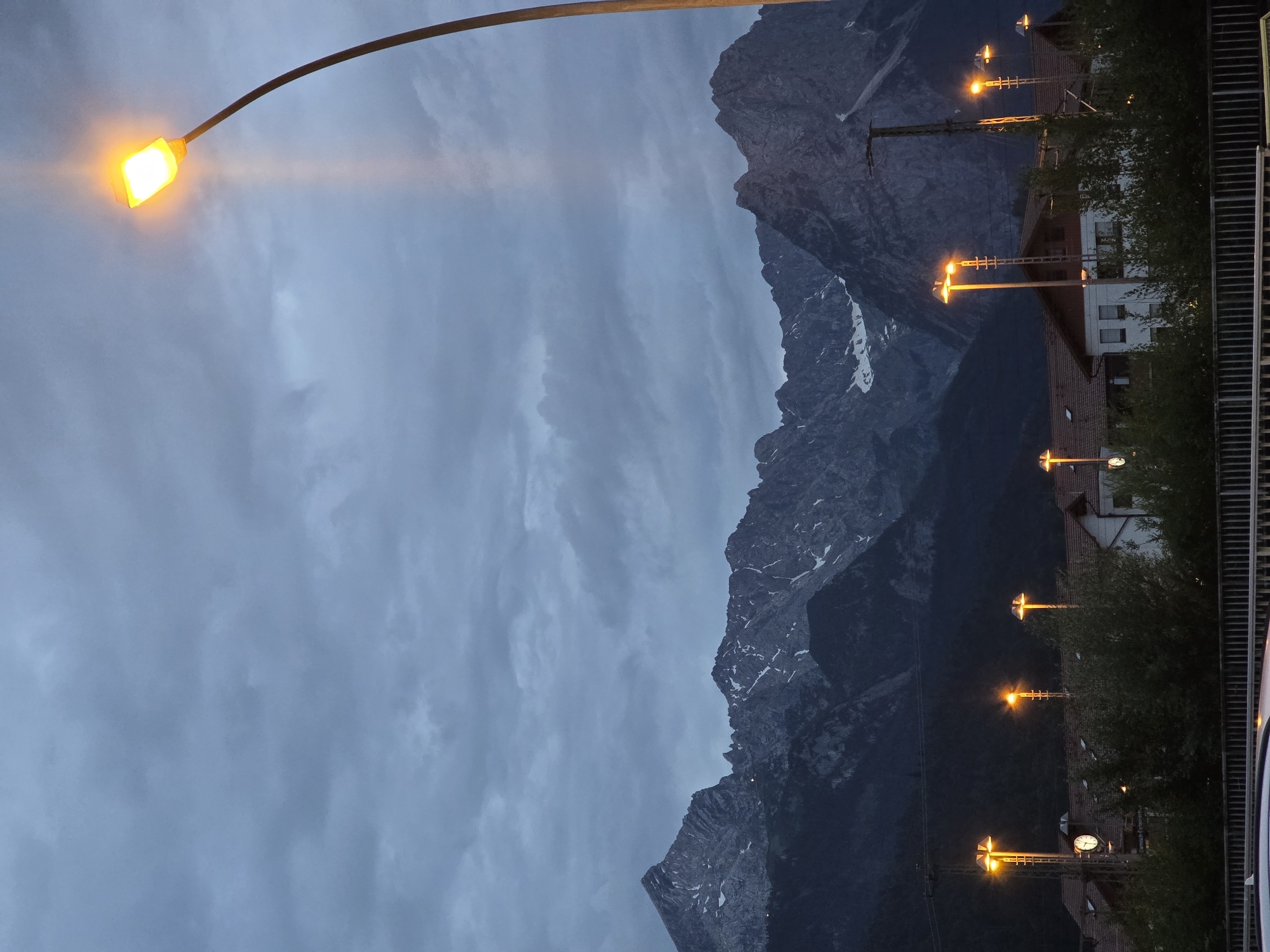 The image depicts a twilight scene in Garmisch-Partenkirchen, Germany, with a dramatic mountain range in the background under a cloudy sky. In the foreground, streetlights illuminate the darkening landscape, casting a warm glow. Several houses with red tiled roofs are visible, interspersed with trees and vegetation. A railway station is implied by the presence of streetlights and possibly a clock tower. The overall atmosphere is serene and slightly melancholic, characteristic of dusk in a mountainous region.