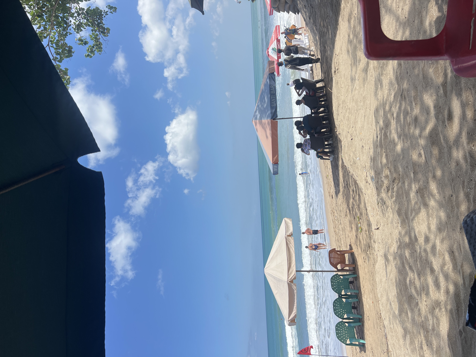 This image depicts a crowded beach scene in Kuta, Indonesia, on a sunny day with a clear blue sky and scattered clouds. The foreground is dominated by the sandy beach and shadows cast by unseen umbrellas and trees, with a hint of a red plastic chair on the right. In the mid-ground, several beach umbrellas provide shade for groups of people seated at tables and chairs. One umbrella is large and beige, with another orange and blue umbrella displaying the text "SANTI BAR". Several individuals are visible walking on the sand or in the shallow ocean. The ocean itself is calm with gentle waves breaking on the shore. A red flag on a pole is planted in the sand on the left side of the frame. The background reveals a tree line and possibly some buildings. The overall atmosphere is one of leisure and activity on a popular tourist beach.