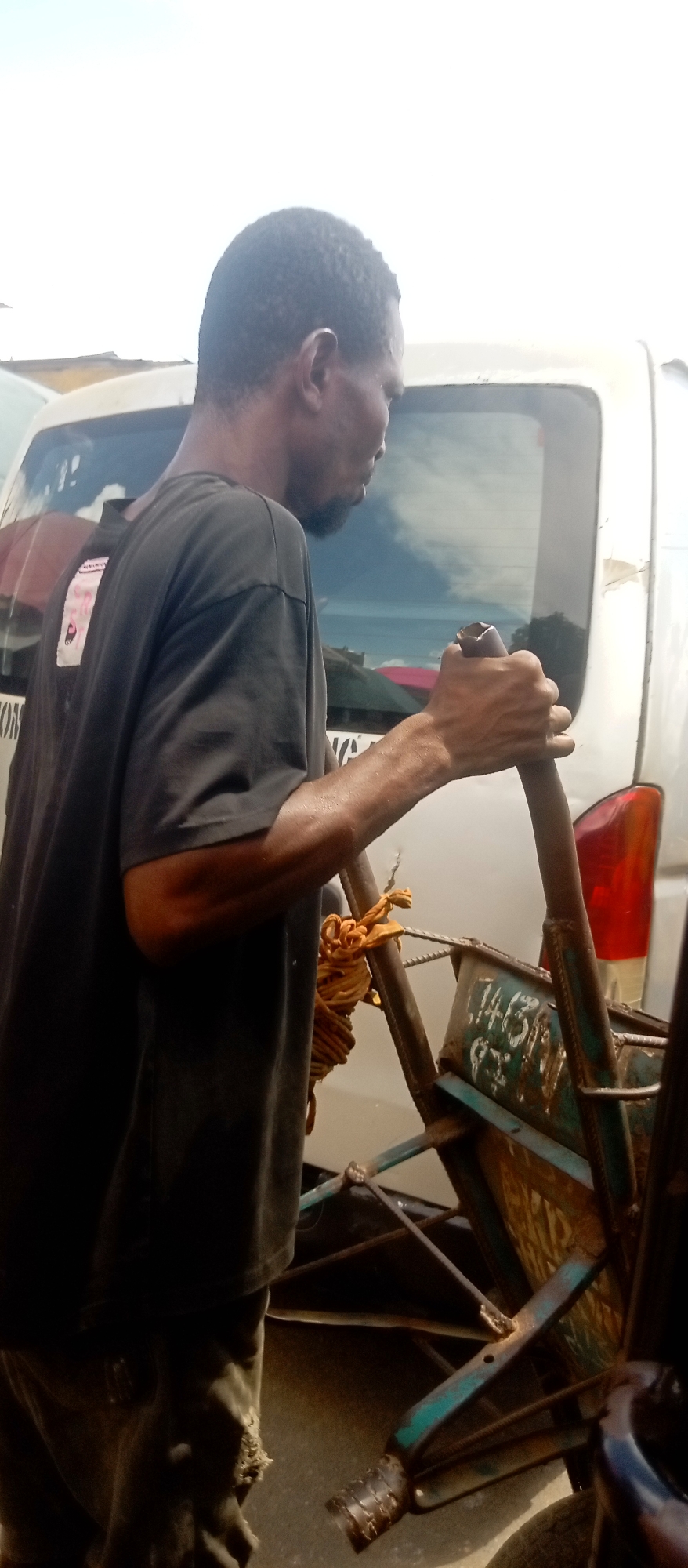A man in a dark t-shirt and worn pants stands next to a rudimentary, metal pushcart in an urban setting. The cart is constructed of welded metal bars and has a container-like section with numbers painted on the side. A rope is tied to the cart. Behind the man and the cart, there is a white van. The location is in the city of Unknown, Unknown. The man appears to be holding onto the handles of the pushcart.
