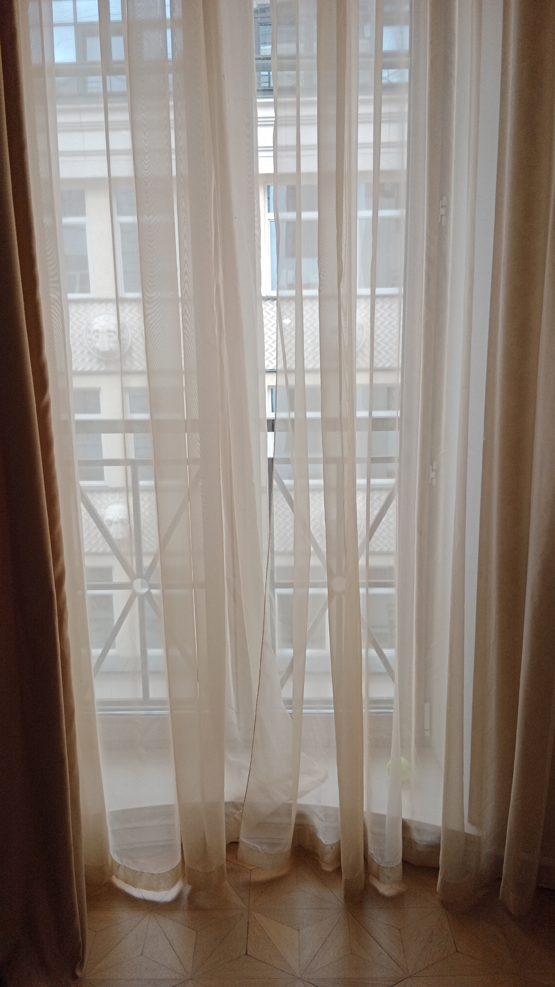 The image shows a view through a window in Moscow, Russia. Sheer curtains obscure the view outside, revealing a building facade with architectural details such as windows and decorative elements. The curtain on the left is a heavier, darker material, creating a contrast with the translucent curtains in the center. The floor has a geometric wood pattern. A faint glimpse of a green ball can be spotted in the bottom right corner, in front of the window.
