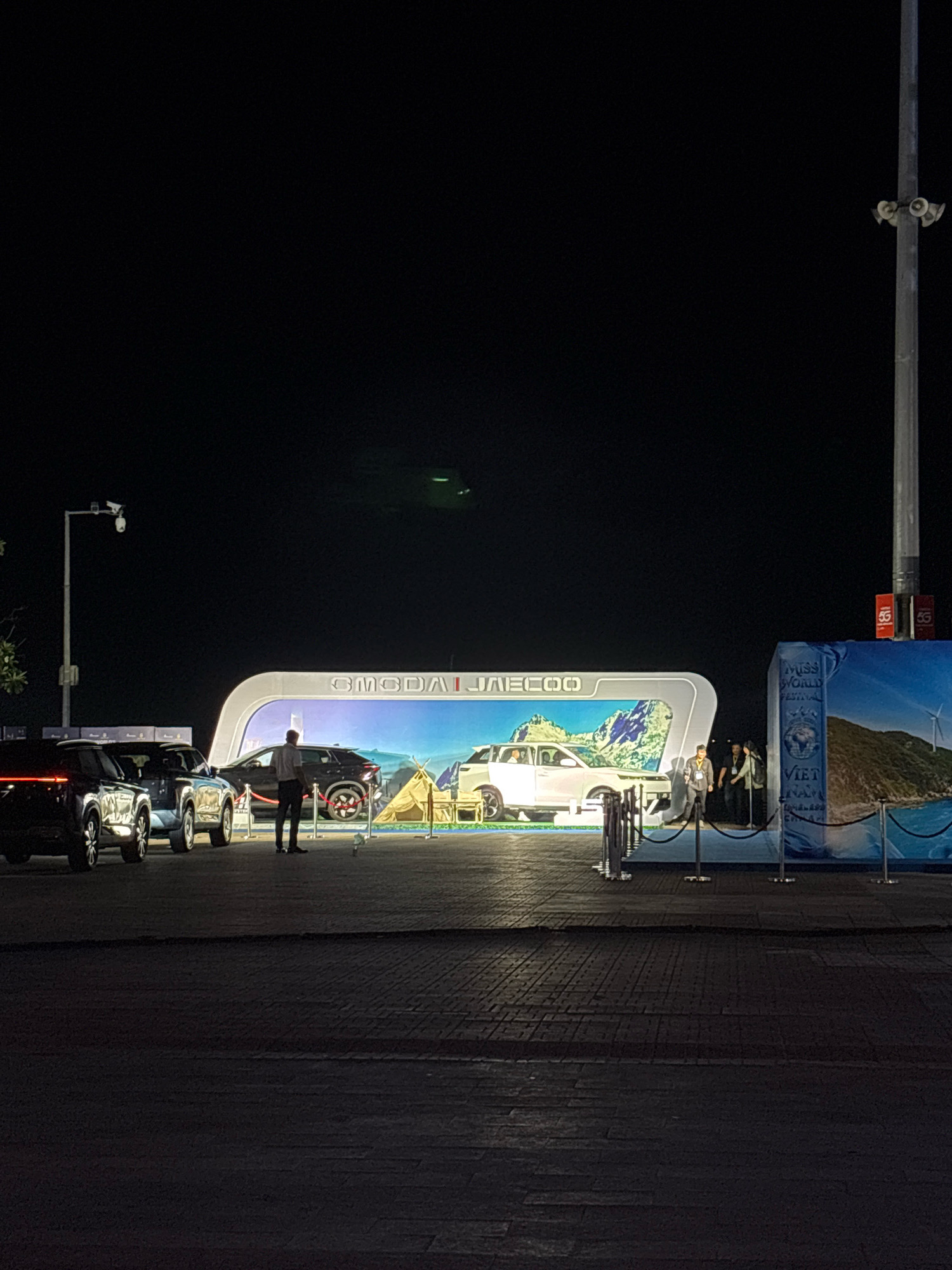 At night in Nha Trang, Vietnam, a car exhibition is set up in an open area. The background features a large, illuminated display with the brand "GMODAI JAECOO" in white, arched lettering. Behind the lettering, a scenic image depicts mountains, a car, and a tent, suggesting an outdoor adventure theme. To the right of the display, a large banner for "MISS WORLD FESTIVAL VIET NAM" is visible, featuring a globe and a coastal landscape with a wind turbine. Several cars are positioned in front of the display, including two dark-colored SUVs on the left and a white car on the right. Three people are present: one individual in a dark shirt and pants stands near the white car, while two other individuals, one in a light-colored jacket and another in a dark outfit, are observed near the banner. The ground is paved with dark rectangular stones. A tall lamppost with attached cameras and speakers stands to the left of the exhibition. The overall lighting is dim, with bright illumination focused on the exhibition area.