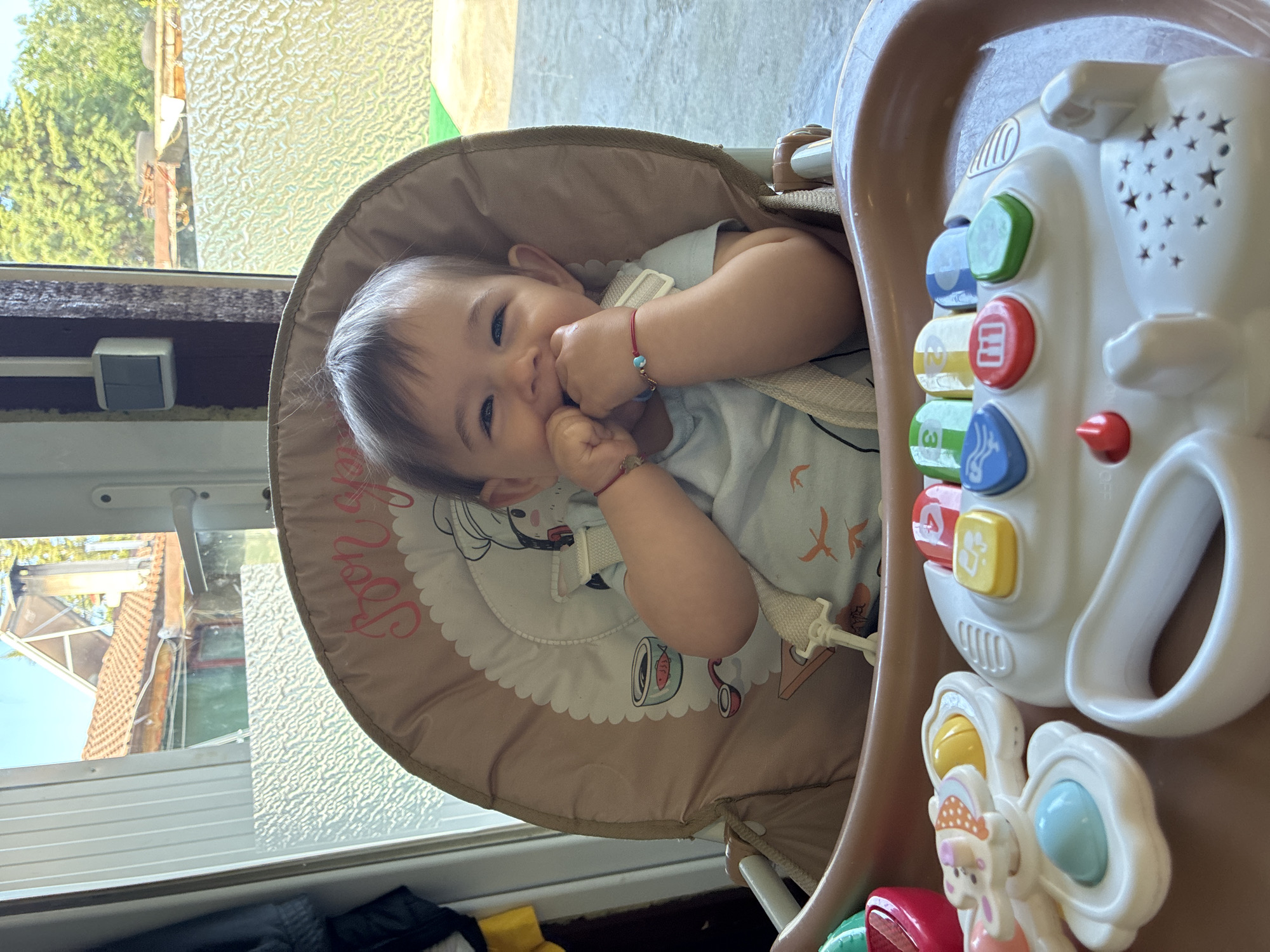 A baby sits in a high chair with an attached toy panel in a room with a window, likely in Varna, Bulgaria. The baby, wearing a light blue shirt, is smiling and holding its hands near its mouth. The high chair is brown with a patterned seat pad. The toy panel attached to the chair has colorful buttons and shapes. Outside the window, there is greenery and the roof of a building visible. The lighting suggests it is daytime.