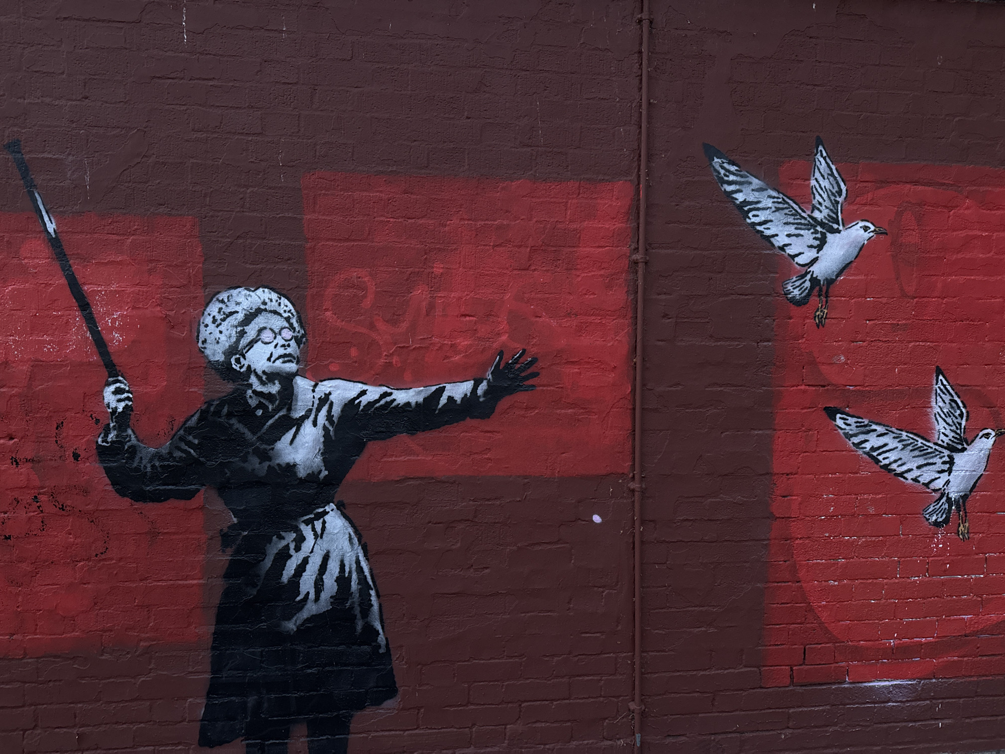 A street art mural is depicted on a textured brick wall, which is painted in varying shades of deep red and dark reddish-brown. The artwork features a grayscale stencil of an elderly woman on the left, characterized by her white hair styled in a bun, glasses, and a long dress. She holds a dark staff or broomstick in her left hand, while her right arm is extended forward, palm open, as if in a gesture of command or release. Her expression, viewed through her glasses, appears focused or stern.
To the right of the woman and above her outstretched hand, two grayscale seagulls are captured in mid-flight, appearing to fly away from her. Their wings display intricate black and white patterns. The background behind the figures includes abstract, geometric shapes, such as rectangles and parts of circles, painted in different shades of red, adding depth to the wall's surface. A thin, dark pipe runs vertically down the right side of the wall. Faint, stylized graffiti, possibly reading "SICK" or "SYCK," is subtly visible in reddish tones on the wall behind the woman's head and outstretched arm. The overall scene evokes a sense of magical interaction between the woman and the birds.