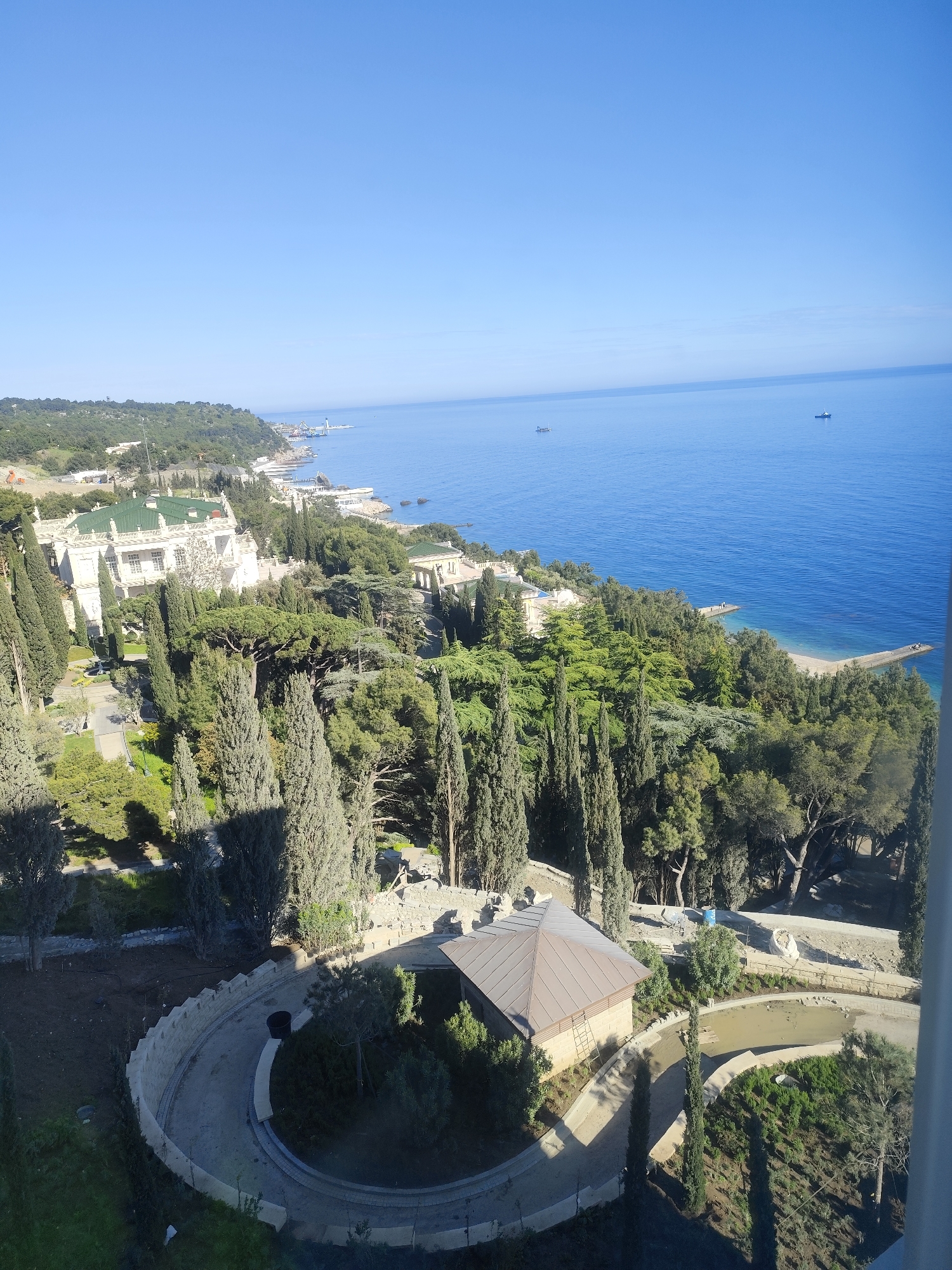 The image presents a high-angle view of a coastal landscape, likely taken from a tall building or viewpoint. The sky is clear and blue, contrasting with the deep blue of the sea which stretches to the horizon. A coastline is visible, featuring a mixture of rocky shores and sandy beaches, dotted with green trees. On the left of the frame, there's a large building with a green roof. In the lower foreground, a smaller structure with a brown roof is visible, surrounded by trees and a circular walkway.