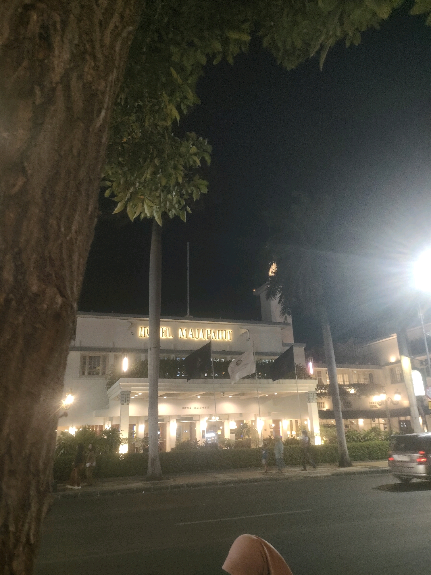 This image captures a nighttime scene in Surabaya, Indonesia, specifically in front of the Hotel Majapahit. The hotel, a historic landmark, is illuminated, showcasing its facade. Several trees are visible in the foreground and midground, with a large tree trunk dominating the left side of the frame. Palm trees are also present. Flags, one white and one black, fly in front of the hotel entrance. People are visible walking on the sidewalk and near the entrance. A car is parked on the street. The street is paved, and a sidewalk separates it from the hotel. The sky is dark, indicating nighttime, and a bright light source, possibly a streetlamp or camera flash, is prominent on the right side of the image, causing lens flare. The overall atmosphere suggests an urban environment at night.
