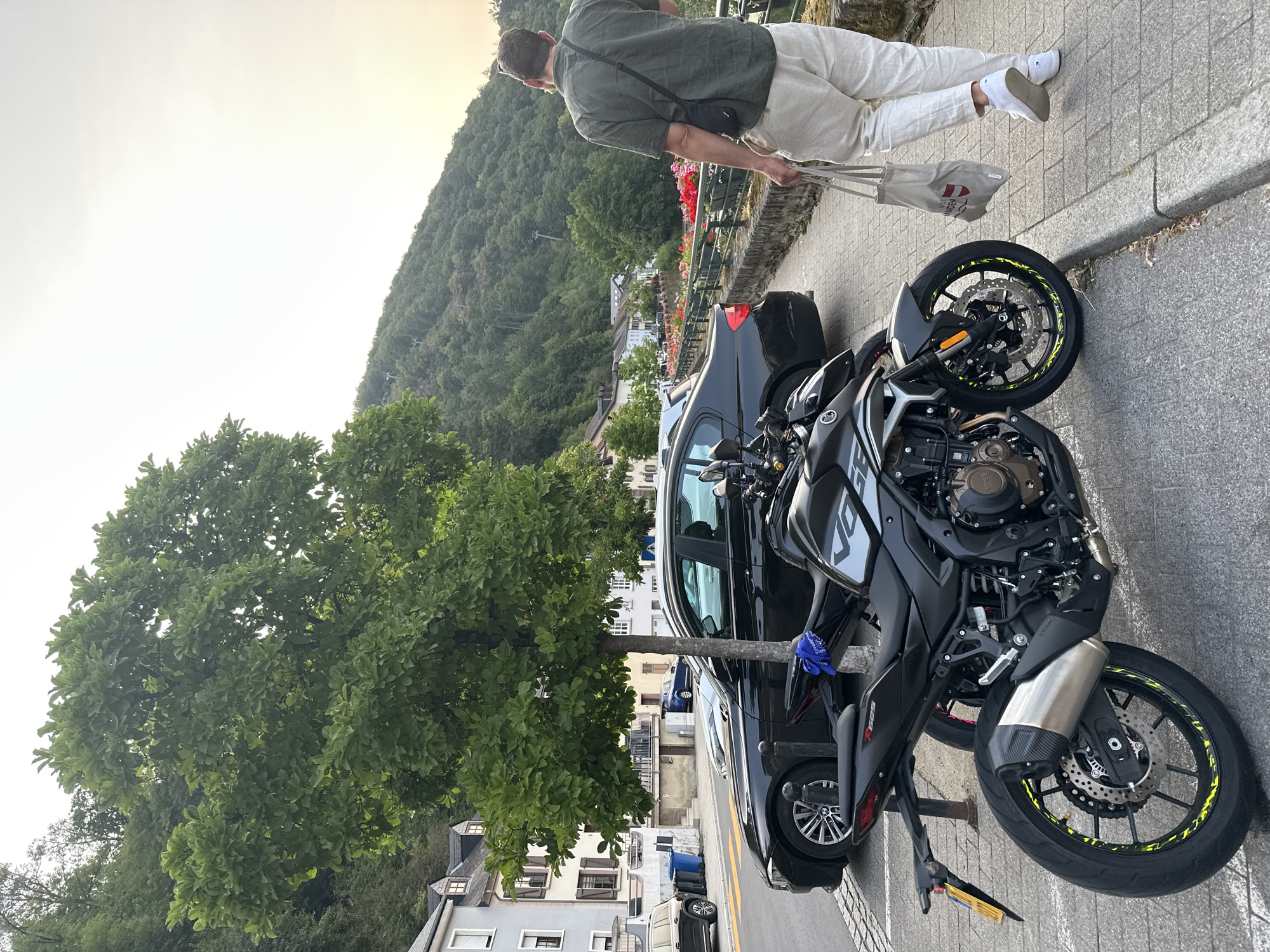 A man walks away from the camera on a cobblestone sidewalk in Vianden, Luxembourg, carrying a tote bag. He is wearing a dark green short-sleeved shirt and light-colored pants, with white sneakers. To his left, parked along the street, are two motorcycles, the closest being black with yellow accents on its wheels. Behind the motorcycles, a black car is parked. Further back, a line of buildings with traditional architecture runs along the street. A lush green tree stands between the street and the buildings. In the background, a steep, tree-covered hillside rises under a hazy sky. The scene appears to be captured during daylight, possibly late afternoon.