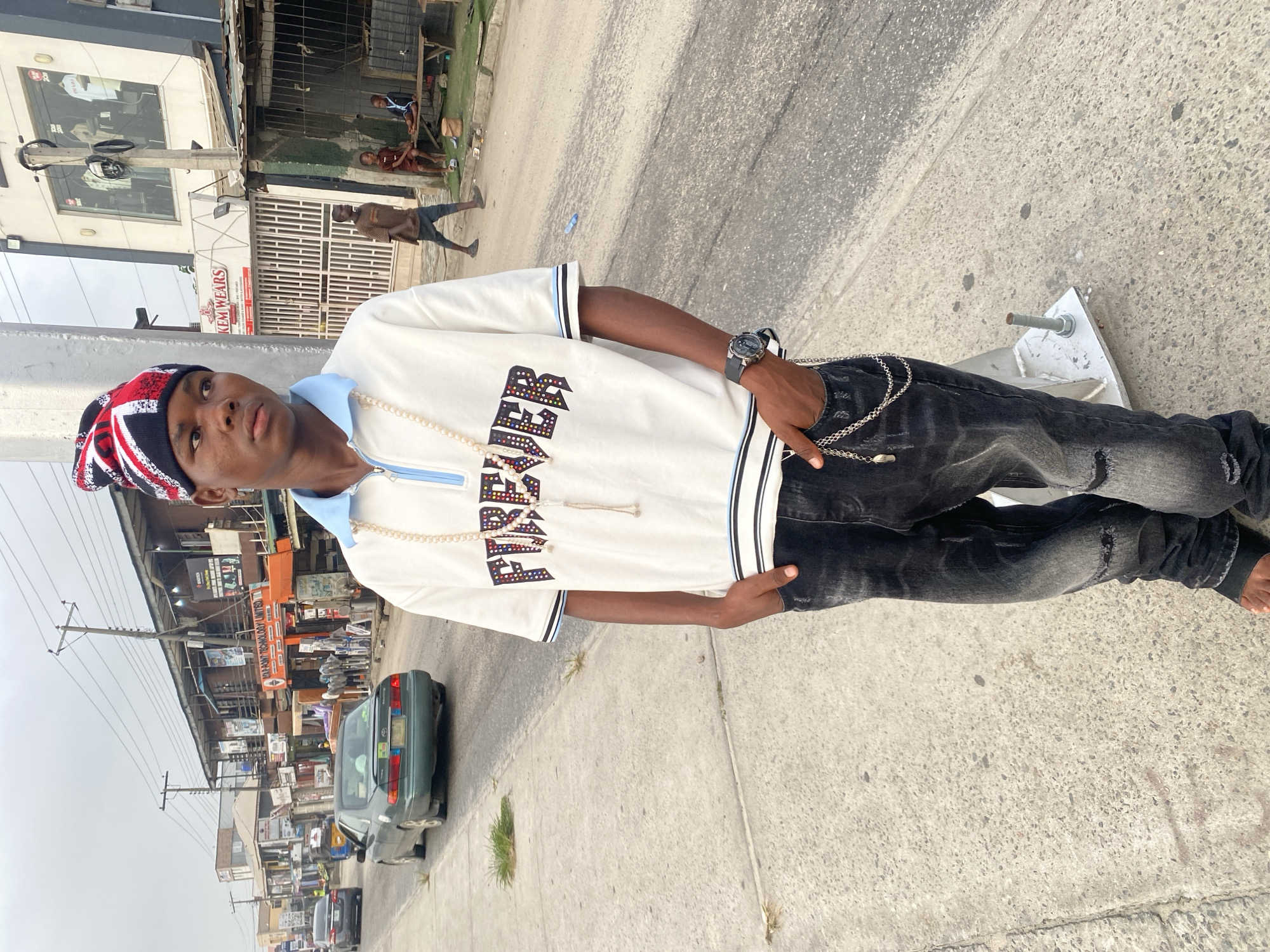 A young man is standing on a concrete sidewalk in what appears to be Ado, Nigeria. He is wearing a white polo shirt with black and white trim, black distressed jeans, and a multi-colored beanie. He has a wristwatch on his left wrist and a silver chain hanging from his jeans. His left hand is in his pocket, and he is looking slightly to the left. The background includes a street scene with various buildings, cars, and pedestrians. There are power lines overhead. Buildings are multi-storied and weathered.