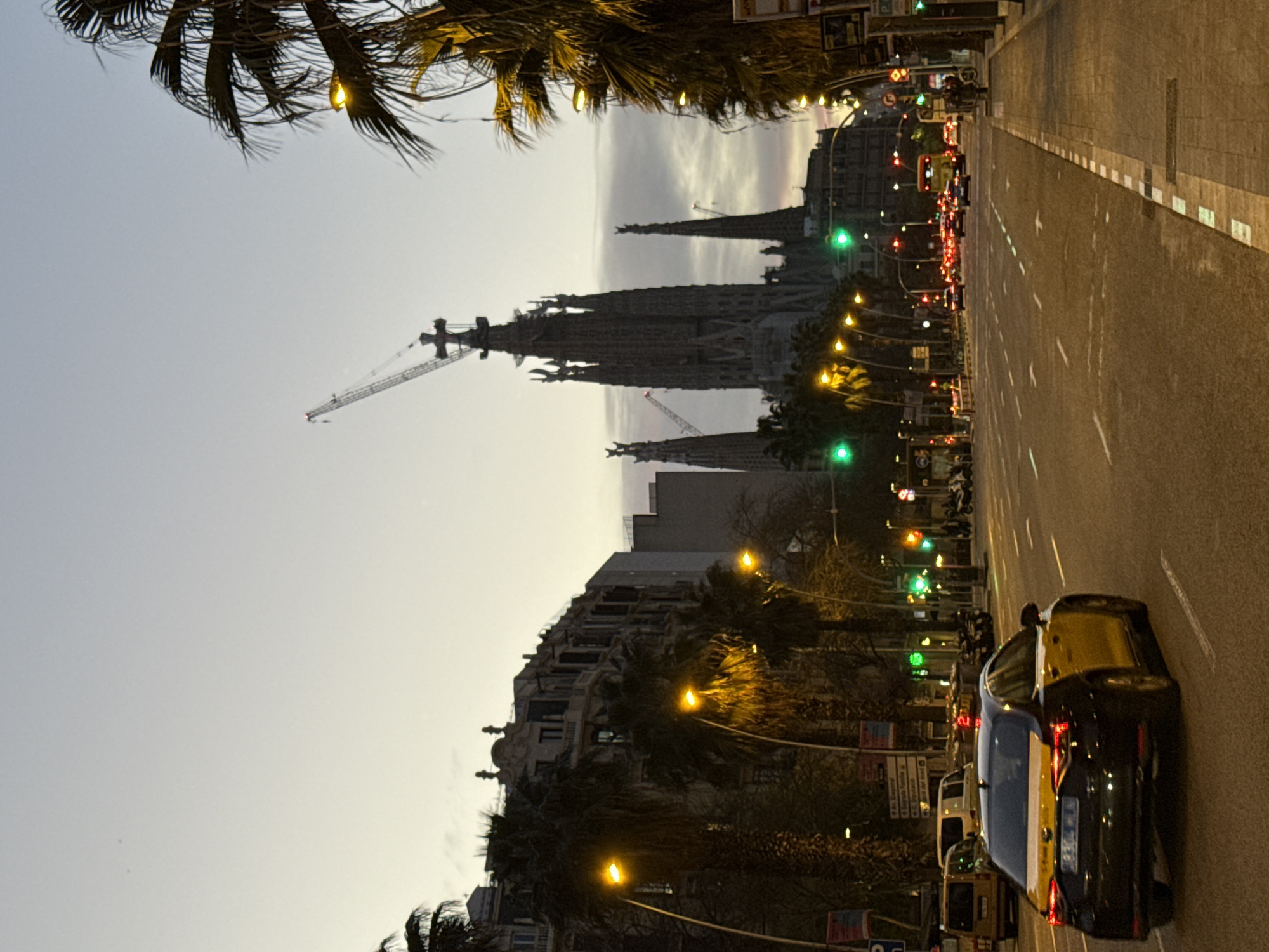 The image shows a street in Barcelona, Spain, at dusk. The Sagrada Familia basilica is visible in the background, partially obscured by palm trees. A construction crane is positioned near the basilica. The street is lined with streetlights, traffic lights, and palm trees. Cars are driving on the road, and some are stopped at a traffic light. The sidewalk is visible on the right side of the image.
