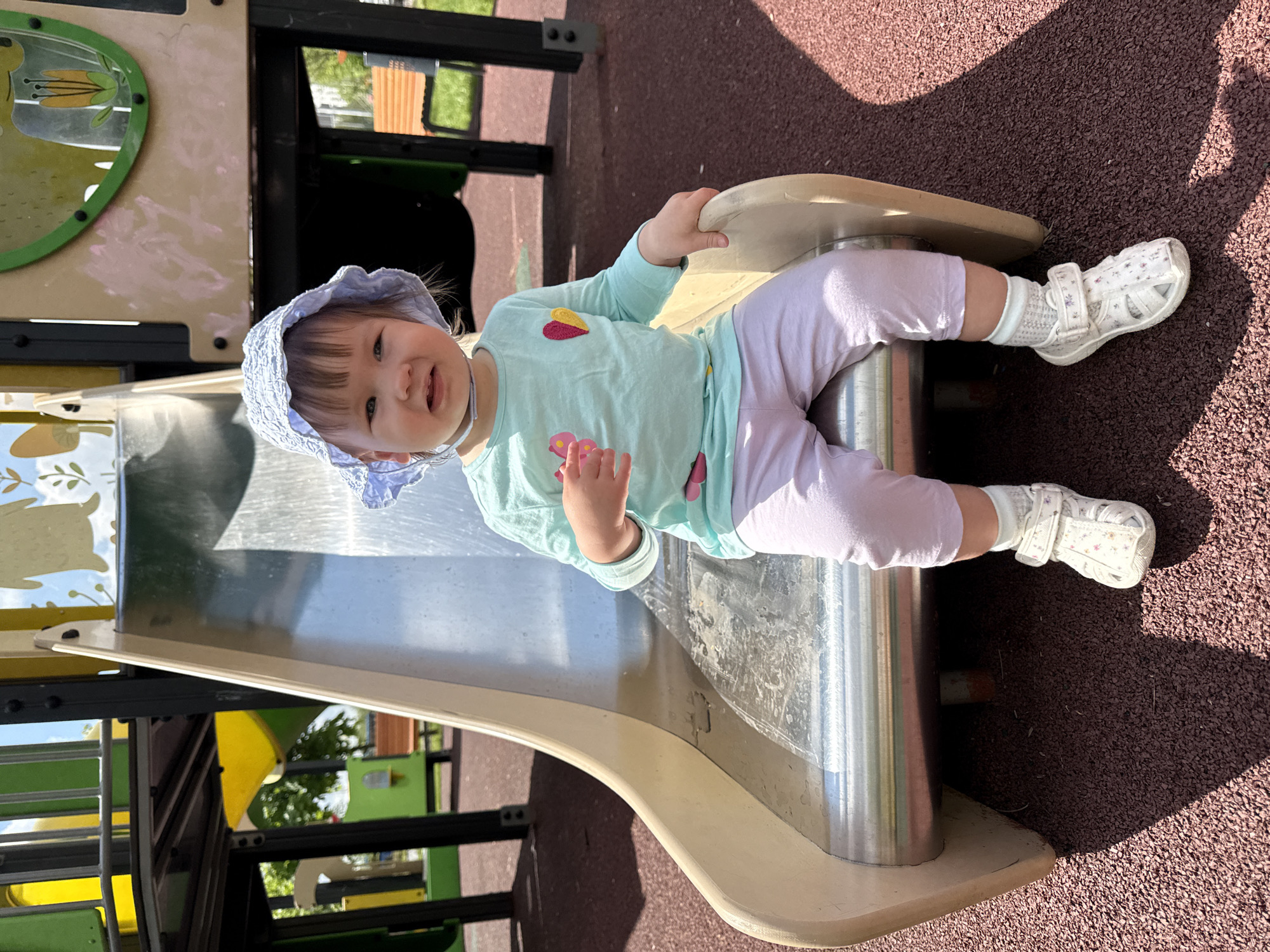 A young child, possibly a toddler, is sitting at the top of a metal and beige playground slide. The child is wearing a light blue patterned bonnet, a long-sleeved light blue shirt with a heart detail, light purple pants, white socks, and white sandals with floral details. The playground surface underneath the slide is a dark red textured material. Parts of the playground structure are visible in the background, including green and yellow elements. The location appears to be an outdoor playground, potentially in Moscow, Russia, based on the vegetation and playground design, but this cannot be definitively confirmed without more information.