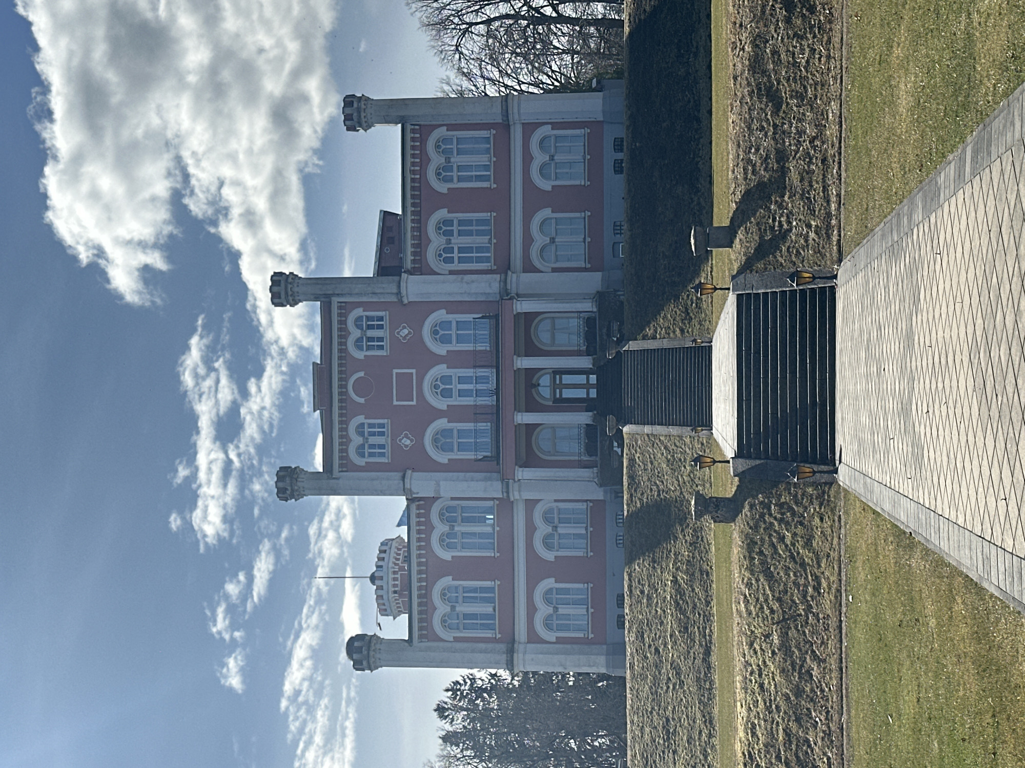 The image captures a vertical view of the Bīriņi Castle in Latvia. The castle has a distinct pink facade with white trim, featuring multiple floors and cylindrical towers topped with crenellations. The sky above is partly cloudy with a blue hue. In the foreground, a pathway leads towards the castle's entrance, flanked by grassy areas and what appears to be a series of steps or a drainage system. The general environment suggests a well-maintained castle grounds.