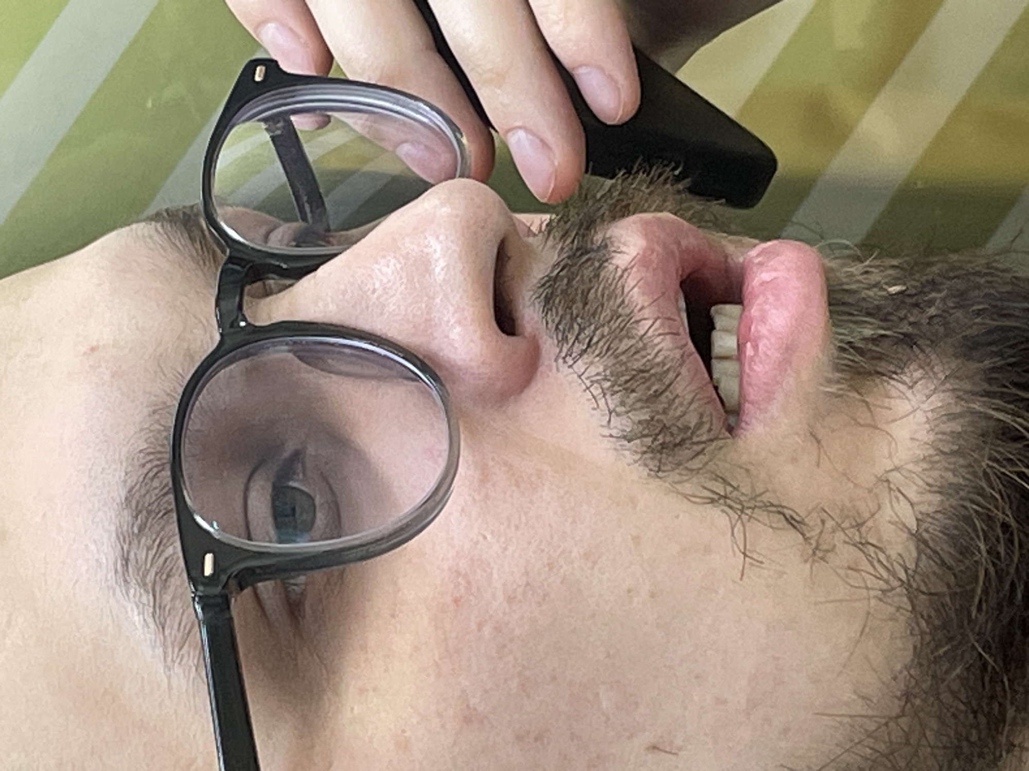 A close-up, high-angle shot depicts a person, likely male, lying down and looking upwards. The person has light skin and visible stubble or short facial hair around the mouth and nose, extending towards the temples. Black-framed glasses are worn, with the left lens partially reflecting a bright light source. The person's mouth is slightly open, revealing some teeth and the inner pink lip. A hand, presumably the person's own, holds a black, rectangular object (possibly a smartphone or remote control) near their forehead, above the glasses. The person's fingers are visible gripping this object. The background features a blurry pattern of diagonal green and white stripes, suggesting an indoor setting such as a wall, blanket, or pillow. No specific time of day, weather, or discernible emotions are evident, and there is no visible text. The provided location context is Krasnoyarsk, Russia, though no specific visual cues within the image confirm this.