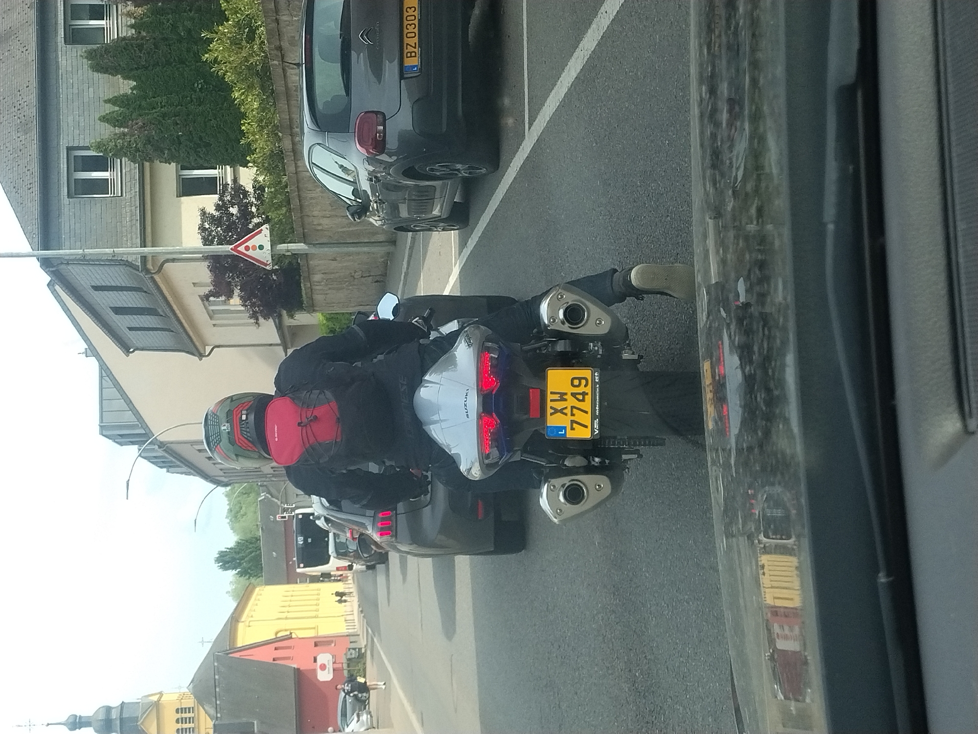 A person is riding a Suzuki motorcycle, bearing license plate XW 7749 from Luxembourg. The motorcycle is traveling on a street in Fentange, Luxembourg. The vehicle is traveling past residential buildings. Parked vehicles are to the right. The photo was taken from another vehicle.