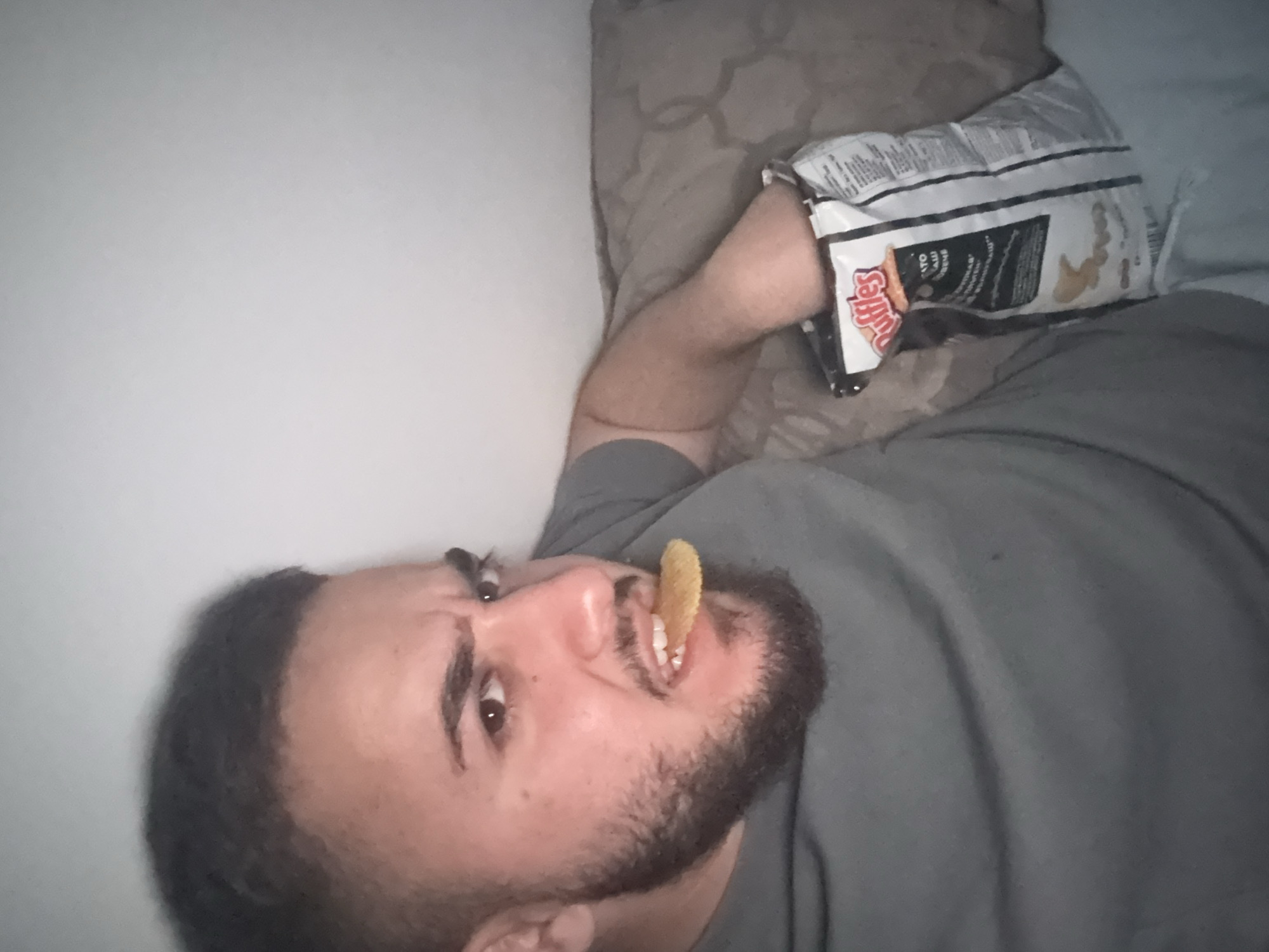 A bearded man lies on his back, holding a bag of Ruffles potato chips open above him. A potato chip is positioned between his teeth. He is wearing a grey t-shirt and appears to be lying on a pillow. The environment consists of a wall and bedding. There is no evidence to suggest this is in Blagoevgrad, Bulgaria.