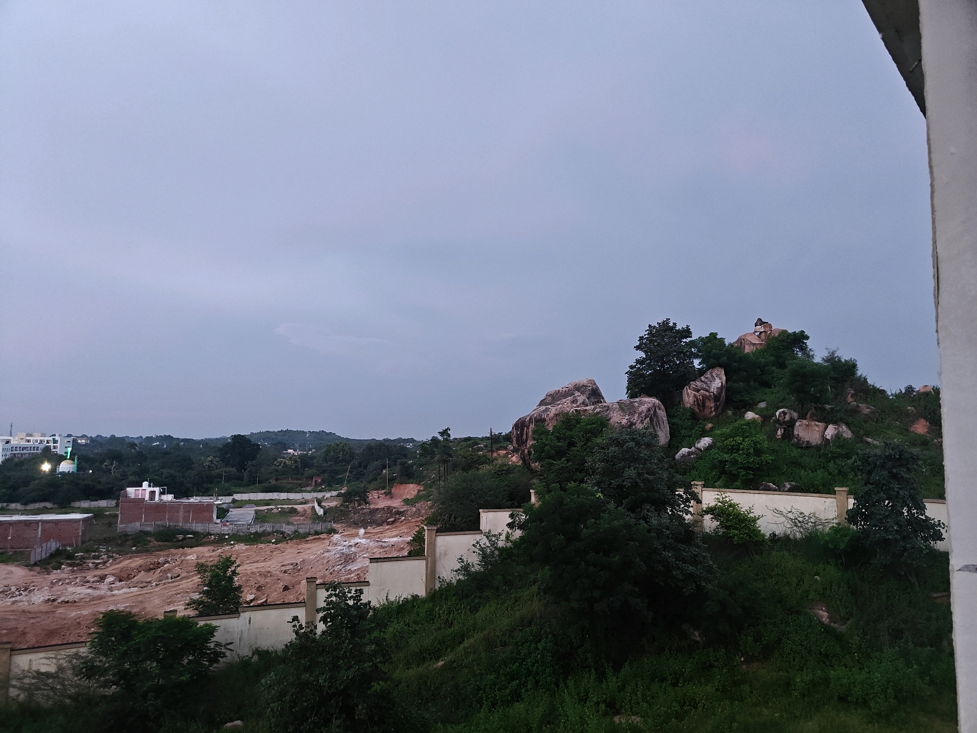 This image depicts a panoramic view of a hilly landscape on the outskirts of Datia, India, likely taken during twilight. The foreground features a wall and lush green vegetation, with some trees obscuring the view of a construction site in the mid-ground. The construction site shows exposed red earth and incomplete brick buildings, suggesting ongoing development. Beyond the construction, the terrain rises into a hill covered with dense green trees and large rock formations. At the summit of the hill, a small structure, possibly a statue or a shrine, is visible. In the distance, a white building with a green dome, likely a mosque, is illuminated, indicating a settled area. The sky is overcast and a pale blue, typical of dusk or dawn. There are no people or animals visible in the frame. The overall scene conveys a mix of natural landscape and urban expansion.