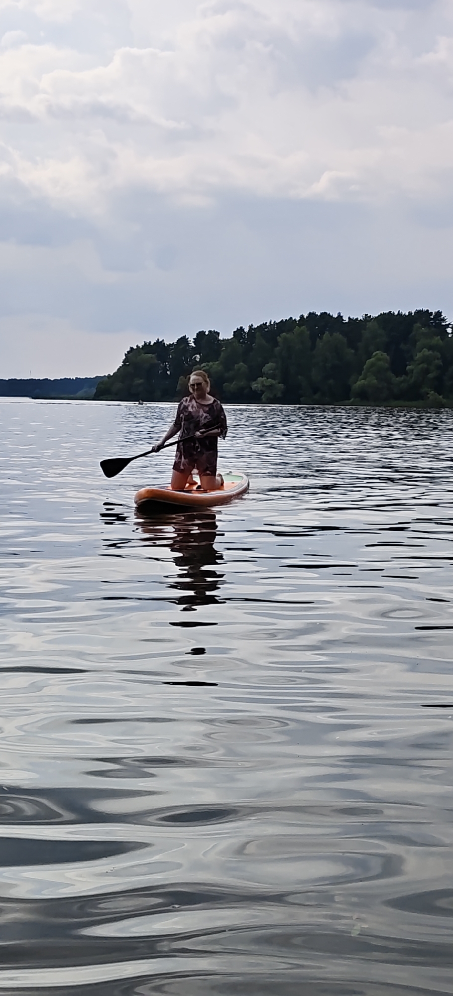 A woman kneels on an orange paddleboard in a body of water, holding a paddle. She is wearing a dark patterned tunic and shorts. Behind her, a dense line of trees forms the shoreline. The sky above is overcast with white and gray clouds. The water is calm with ripples reflecting the sky and trees. The scene appears to be a natural setting, likely a lake or river, located in a rural area. No specific city or landmark is identifiable within the image.
