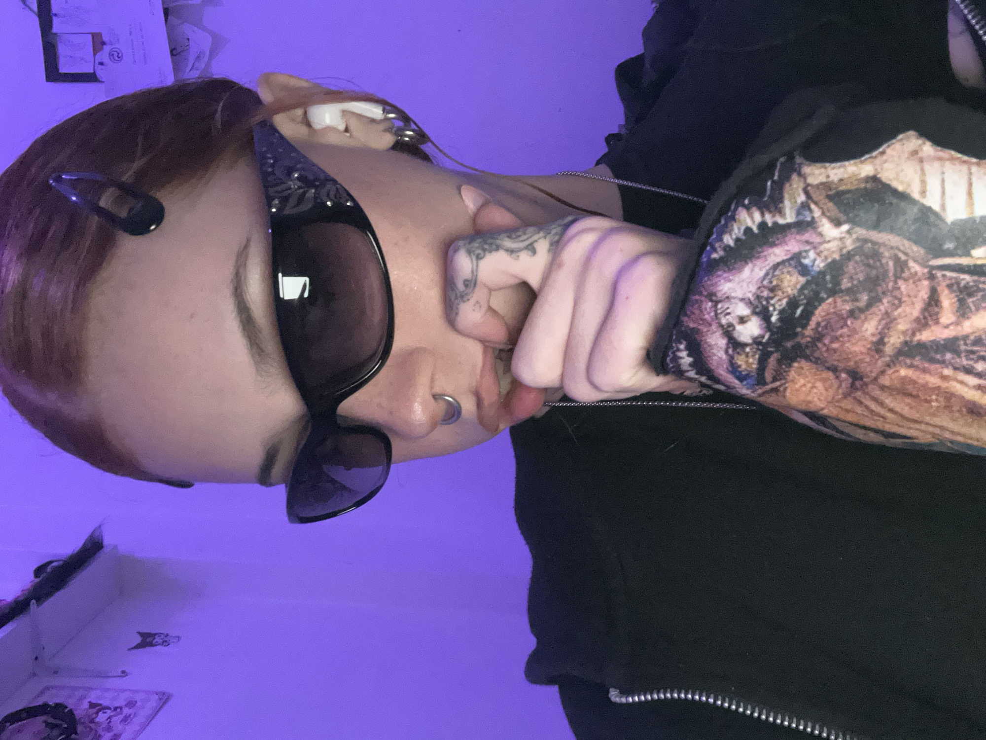 An angled, close-up shot captures a person with reddish-brown hair, featuring black snap clips. They are wearing dark, possibly reflective, sunglasses with what appears to be an ornate or patterned frame. The individual displays multiple body modifications, including a septum piercing and intricate tattoos on their right hand and forearm, featuring swirling, organic patterns.
The person is dressed in a dark, likely black, hooded jacket with a silver zipper, worn over a t-shirt that reveals a complex, multi-colored graphic print on the sleeve/shoulder area. This print depicts an artistic, possibly macabre or fantasy-themed scene with various figures and textures. Their left hand is raised, with fingers curled and touching their lower face/mouth area. A thin silver chain is visible, likely a necklace or part of the clothing.
The setting appears to be indoors, dominated by a vibrant, artificial purple light emanating from the background, suggesting mood lighting, possibly in the evening or night. In the upper background, some papers are loosely affixed to a surface, and a small framed item or drawing can be seen. The image provides no specific visual cues for the time of day or weather beyond the artificial lighting. No discernible text is visible within the frame. This photo was taken in Buenos Aires, Argentina.