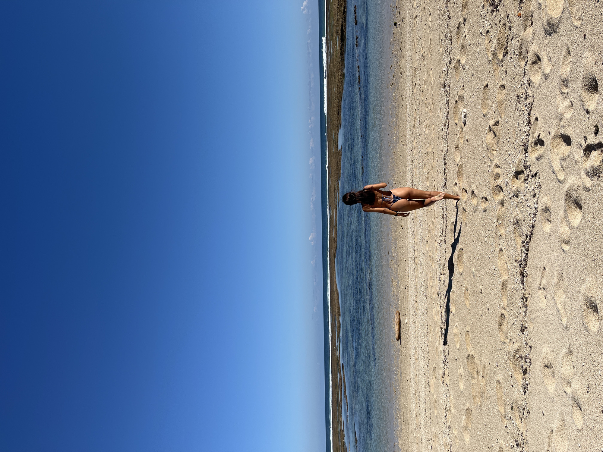 This image, when correctly oriented, portrays a serene beach scene bathed in bright sunlight in Kuta Selatan, Indonesia. The dominant subject is a person, appearing to be a woman with long dark hair, lying on her stomach on the sandy shore, facing away from the viewer. She is dressed in a patterned bikini.
The foreground consists of light-colored sand, marked with numerous footprints and scattered small rocks. A small piece of driftwood is also visible on the sand. To the right of the person, the clear, shallow ocean water meets the shore, revealing a rocky or coral-strewn seabed near the waterline. Further out, small waves can be seen breaking along the distant horizon.
The upper portion of the image features a vast, clear blue sky, indicating excellent weather conditions. The strong sunlight casts distinct shadows, suggesting the time of day is around midday or early afternoon. The overall impression is one of peaceful relaxation on a sunny, tropical beach. No text is visible in the image.