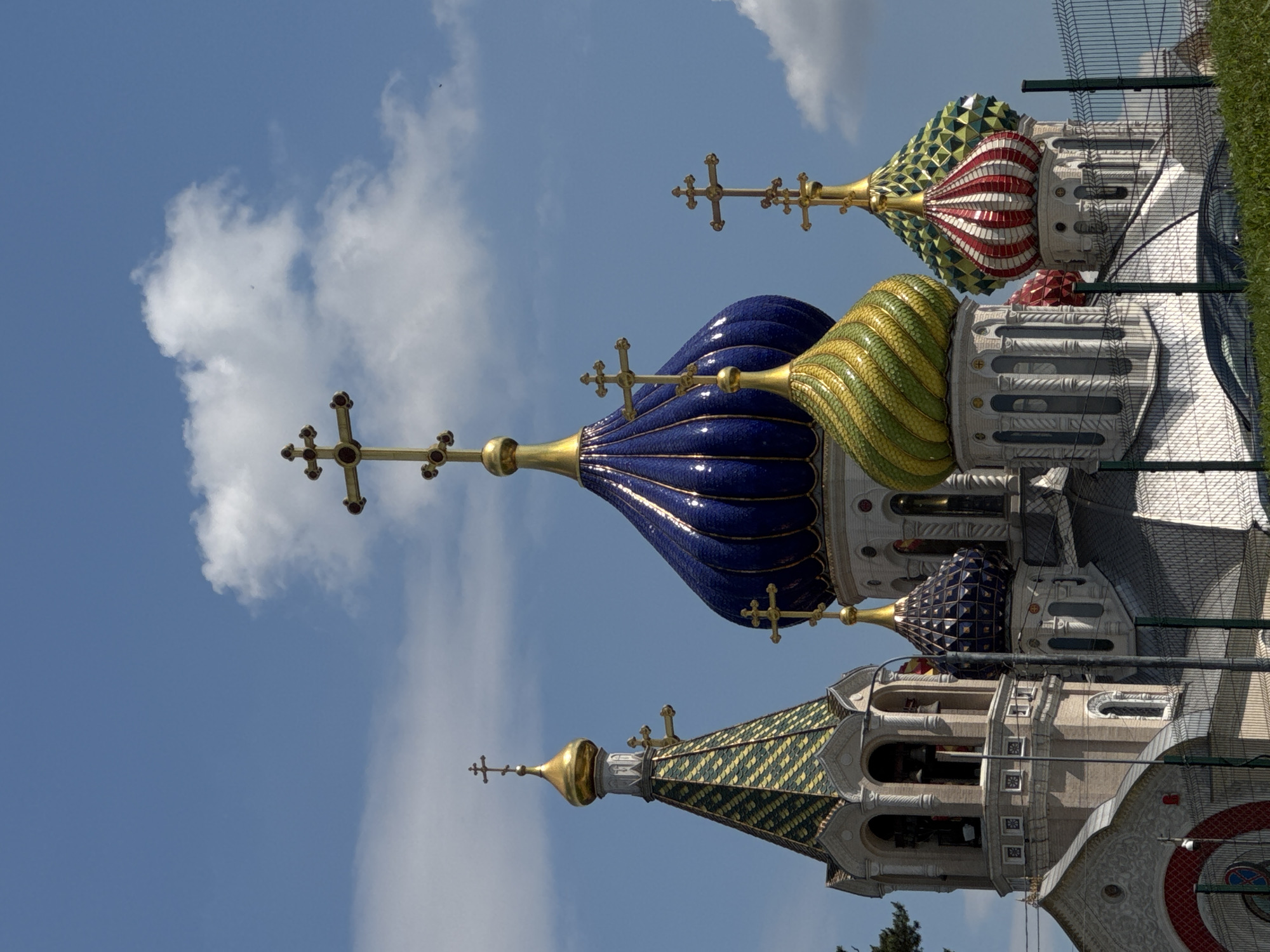 The image captures a vibrant and ornate collection of onion domes, characteristic of Russian Orthodox architecture, set against a clear blue sky dotted with fluffy white clouds. The primary subjects are the colorful domed spires of what appears to be a church or cathedral.
A prominent large dome dominates the center, featuring a deep blue, mosaic-like surface that reflects the light. It is crowned with a gleaming golden Orthodox cross. To its right, another dome showcases a distinctive green and yellow striped pattern, also topped with a golden cross. Further right, a striking red and white striped dome is partially visible, alongside a smaller, darker blue dome with a geometric pattern, and a green and yellow checkered dome that seems to be part of a larger bell tower structure. Each dome is surmounted by a golden Orthodox cross.
Below the colorful domes, intricate architectural details of the building are discernible, including white and gray stone facades with arched openings and decorative elements. The bright conditions indicate it is daytime, likely midday or afternoon, under sunny weather. In the background, particularly on the right side, a green chain-link fence is visible, suggesting the area might be enclosed or a construction site, with some green foliage beyond. There are no people or discernible activities taking place, and no visible text in the image. The scene conveys a sense of architectural grandeur and spiritual beauty.