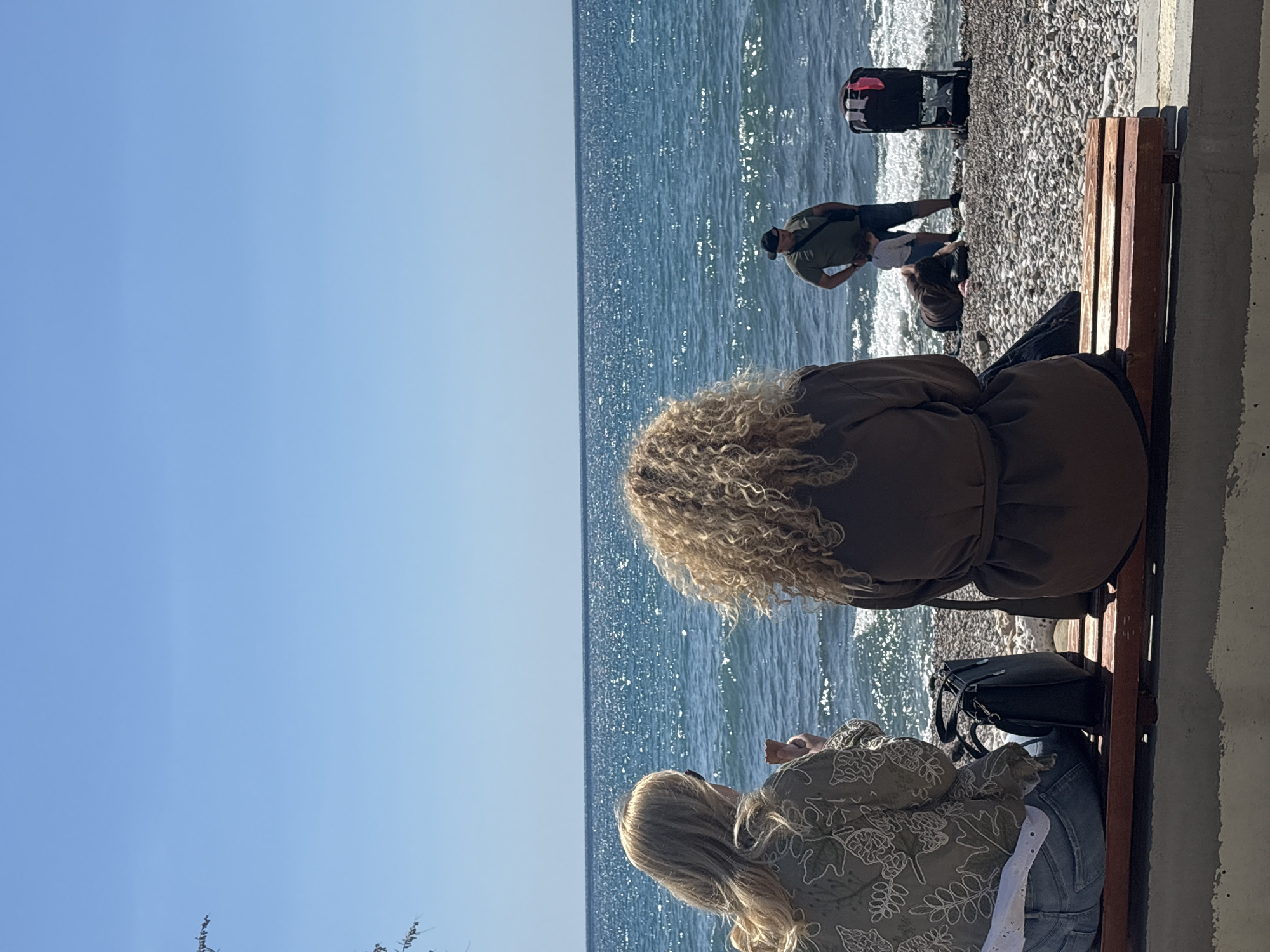 Two women are sitting on a wooden bench, with their backs to the viewer. The woman on the right has curly blonde hair, and is wearing a brown dress with a belt. The woman on the left has straight blonde hair and is wearing a light green flowered shirt and blue jeans. To their left, the Adriatic Sea shimmers under a clear blue sky. Further away, a man wearing a green shirt and a black hat is looking down toward a smaller individual on the stony beach, with a black stroller behind them. A black handbag is placed on the bench next to the seated women. The scene is located in Bar, Montenegro, next to a promenade and beach. A concrete wall is on the right-hand side of the bench.