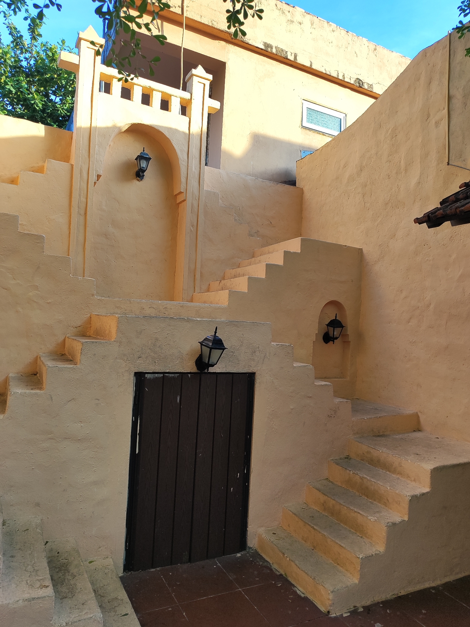 The image depicts an outdoor staircase with a rustic, weathered aesthetic, likely in an older part of Đà Nẵng, Vietnam. The walls are painted a warm, ochre hue, textured and showing signs of age. Sunlight casts strong shadows, highlighting the architectural details.
The staircase itself is made of concrete, with wide, shallow steps that appear worn. The risers are painted the same ochre as the walls, contrasting with the lighter, grayer treads. On the wall to the left of the staircase, there is a dark wooden door with vertical planks. Above the door, a black, traditional-style outdoor lamp is mounted within an arched recess.
Further up the staircase, on a landing, another identical lamp is mounted on the wall. To the right, the staircase continues upwards, with more steps leading to an unseen upper level. A small, arched niche is visible on this section of the wall, also containing a black lamp.
In the background, the building exhibits more of the same ochre plaster. A small, rectangular window with a patterned glass pane is visible high on the wall. Green foliage from a tree hangs into the frame from the top left, and the sky above is a clear blue. The ground in the foreground appears to be tiled with dark red or brown square tiles. No people are visible in the image. The scene evokes a sense of history and a quiet, perhaps less-trafficked, corner of the city.