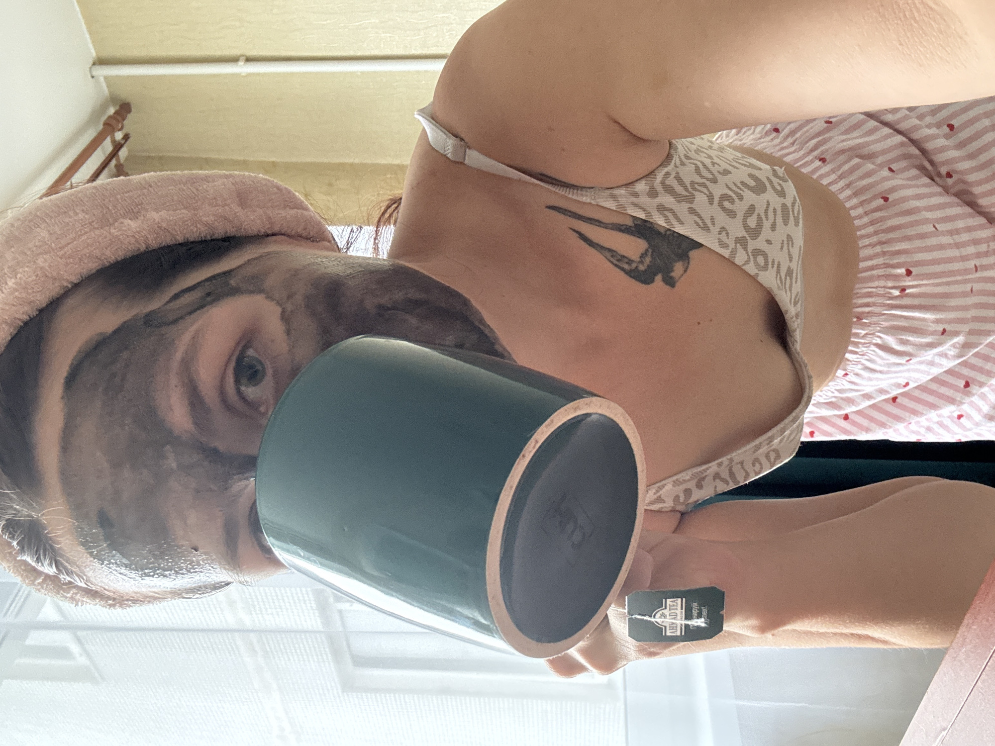A woman is shown from the chest up, drinking from a dark green mug. She has a face mask on and a pink towel wrapped around her head. She is wearing a light-colored bra with a leopard print and pink striped pajama bottoms with small red hearts. A black tea bag with the brand "AHMAD TEA" is hanging from the mug. A tattoo of a bird in flight is visible on her upper chest. The background appears to be indoors, with a window on the left showing a view of a building with multiple windows. The lighting suggests it is daytime. The scene is likely within a residential setting in Naberezhnye Chelny, Russia, given the context.