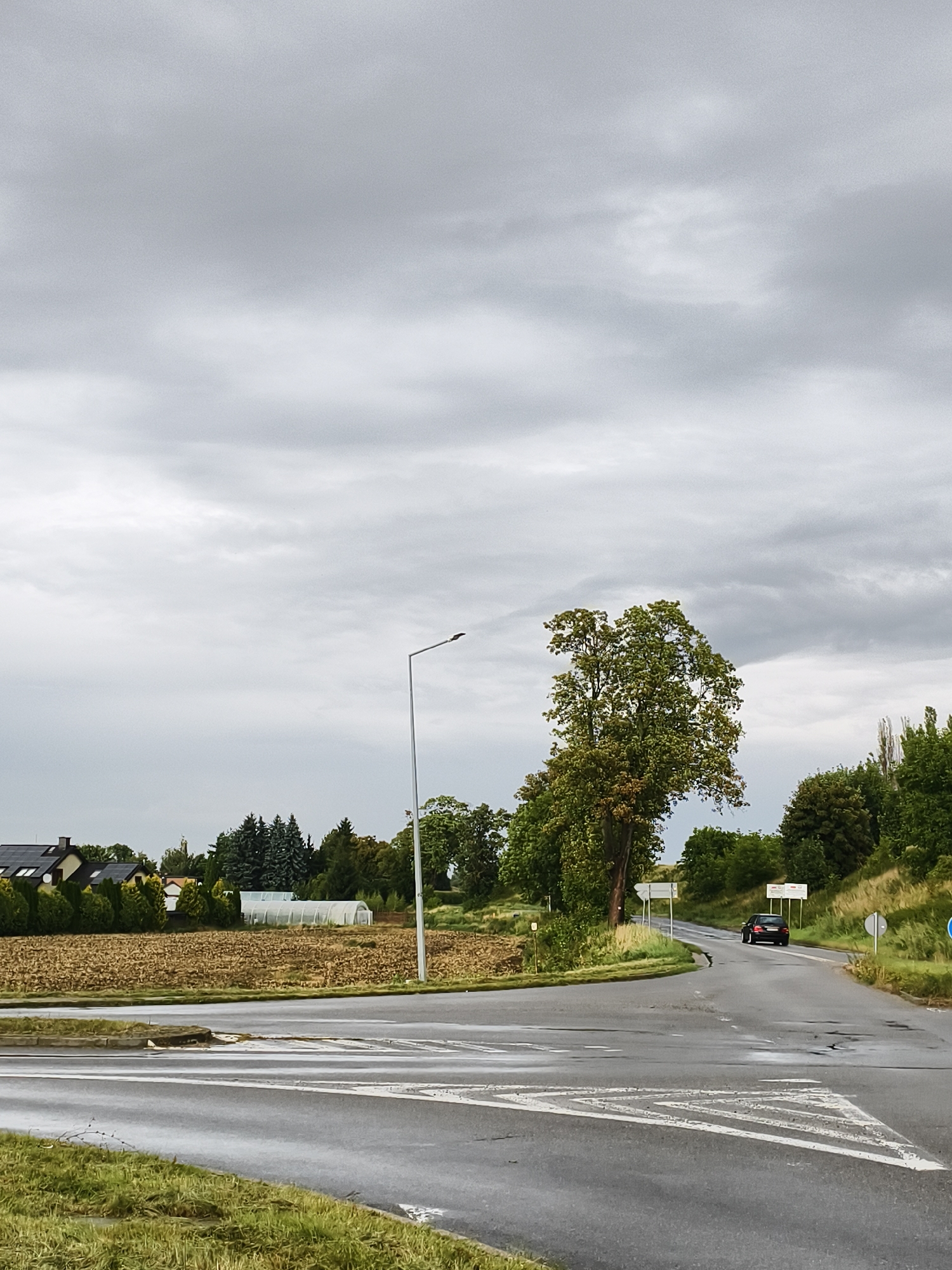 This is a photograph of a rural intersection on a cloudy day. The road is wet, indicating recent rain. There is a black car driving away from the camera on the right side of the road. On the left side of the road, there is a brown field and some houses with solar panels on their roofs. Further in the background, there are trees and a greenhouse. A street lamp is visible in the middle of the frame. The location is the intersection of ulica Jana Pawła II and ulica Krótka in Tczew, Poland.