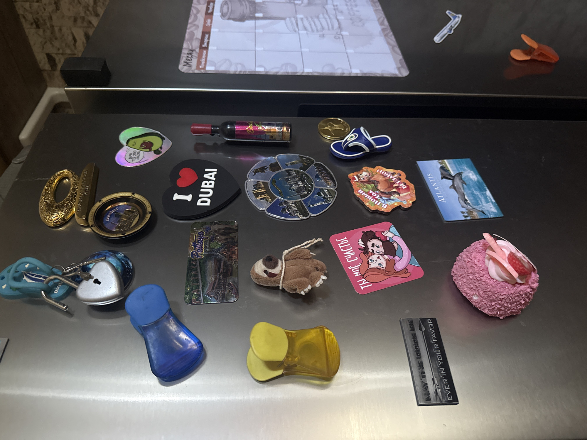 The image shows a close-up of a refrigerator door, adorned with various magnets and small objects. The refrigerator has a stainless steel finish.
On the upper left, there is a blue plastic object, possibly a bag clip or a novelty item, with a shiny metallic finish. Below it is a bright yellow, translucent plastic object shaped like a pair of flip-flops.
Further down on the left, a rectangular black magnet with silver lettering reads "MAY THIS GLOBE BE EVER IN YOUR FAVOR". To the right of this, a round, pink, textured object resembling a dessert or a cake, decorated with pink frosting and a strawberry slice, is attached to the door.
In the center of the image, there are several souvenir-like magnets. One depicts a heart with an avocado inside, with text in an unfamiliar script. Below this is a prominent black magnet shaped like a heart with the words "I ♡ DUBAI" in white letters. To the left of the "I ♡ DUBAI" magnet is a circular magnet with multiple images arranged around a central motif, possibly depicting landmarks. Adjacent to this is a small, miniature bottle with a red cap, featuring colorful labels.
Above the "I ♡ DUBAI" magnet, there's a silver heart-shaped locket with a keyhole, hanging from a blue flip-flop shaped keychain with a metal ring and a key. To the left of this, a golden oval ornament with ornate detailing is attached.
On the lower right side, there is a magnetic calendar with the heading "Месяц" (Month) and days of the week listed in Russian. To the left of the calendar is a magnet depicting two dolphins leaping out of the water in front of a tropical beach scene, with the word "ATLANTIS" visible. A small, blue and white flip-flop shaped magnet is positioned above the Atlantis magnet.
Near the top of the right side, a miniature airplane is attached to the door. At the bottom right, an orange object, possibly a clip, is attached.
The overall environment appears to be a domestic kitchen setting. The lighting suggests an indoor environment, likely lit by artificial light or ambient room light. There are no people visible in the image.