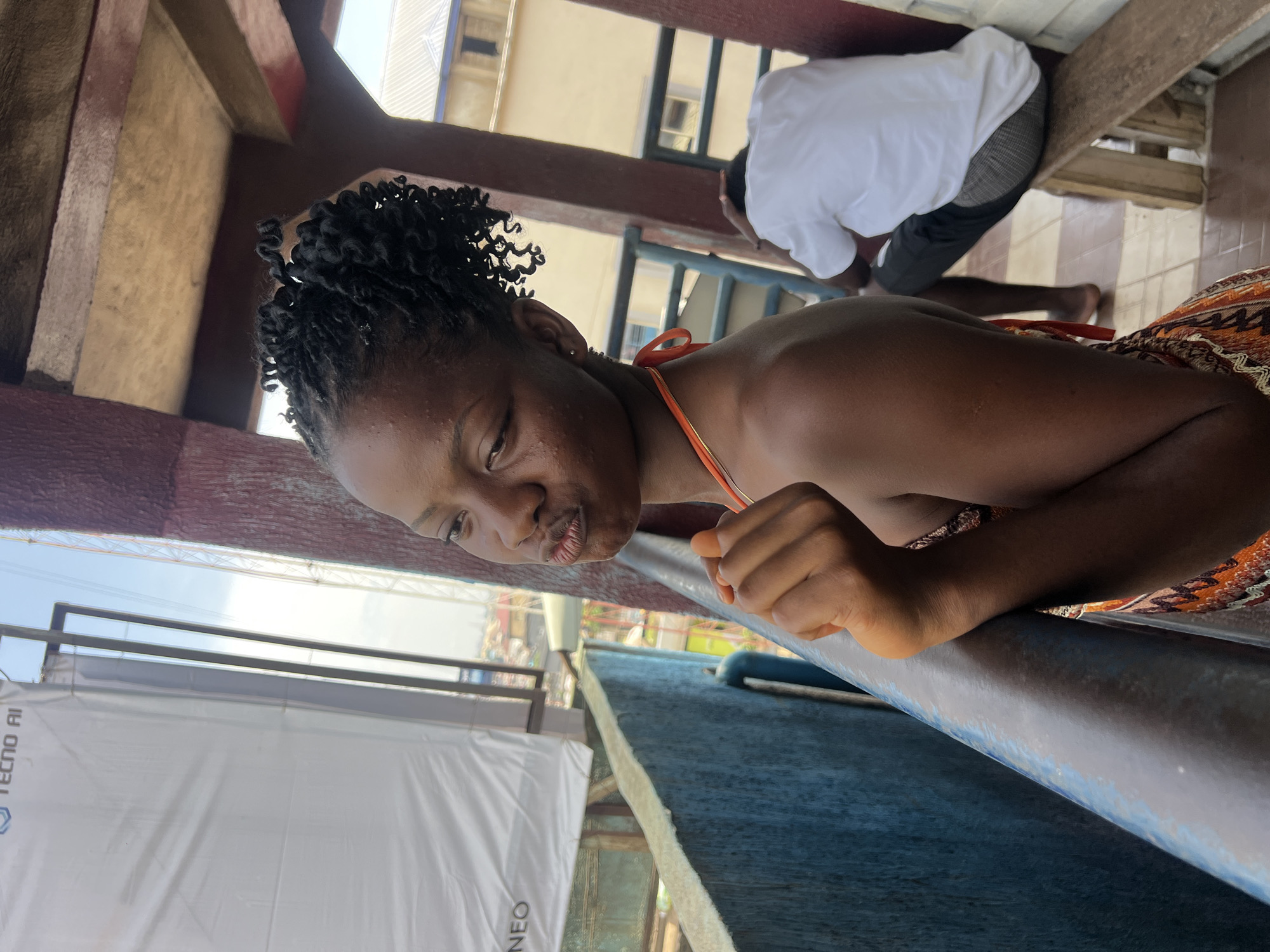 The image shows a young Black woman with curly black hair, wearing an orange string bikini top and a patterned skirt. She is leaning on a railing and looking down with pursed lips. Her skin is dark and appears to be sweating. In the background, another person wearing a white shirt and plaid shorts is kneeling on the ground. The scene appears to be outdoors, possibly on a balcony or porch, with wooden structures and a tiled floor visible. A large white tarp with "TECNO AI" printed on it is in the lower left portion of the image, suggesting it might be near an event or display. The lighting suggests it is daytime, and the overall atmosphere seems casual and relaxed. The location is identified as Nkpor, Nigeria.