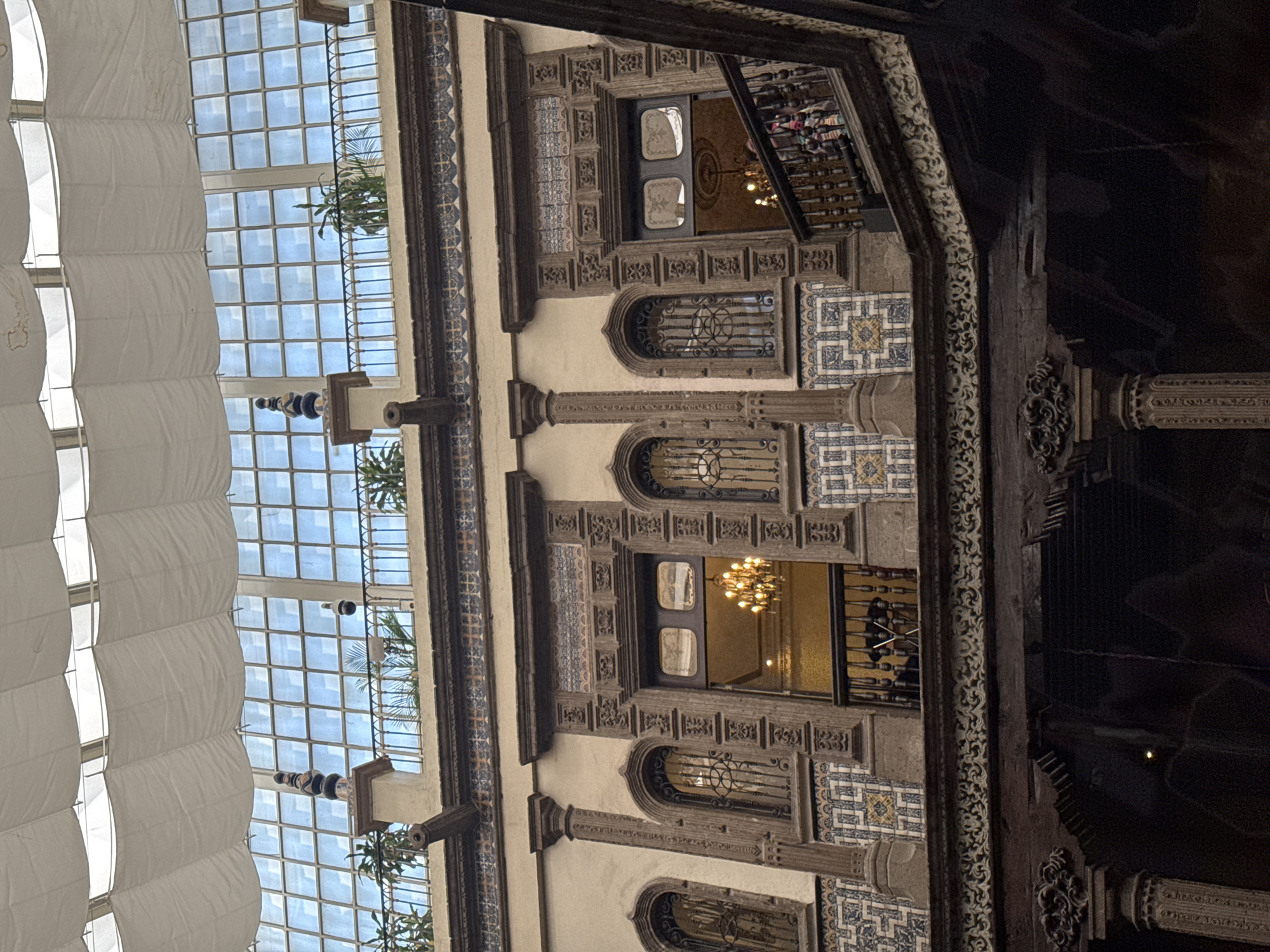 The image shows a view of the Gran Hotel Ciudad de México interior, located in Mexico City, Mexico. The ornate building interior features decorative stonework with blue and white tiles, archways, and balconies. A glass-covered ceiling with white draperies hanging beneath it provides natural light. Chandeliers are visible through the openings on different floors, and figures can be seen in the background. The architectural style is intricate and reflects historic design elements.