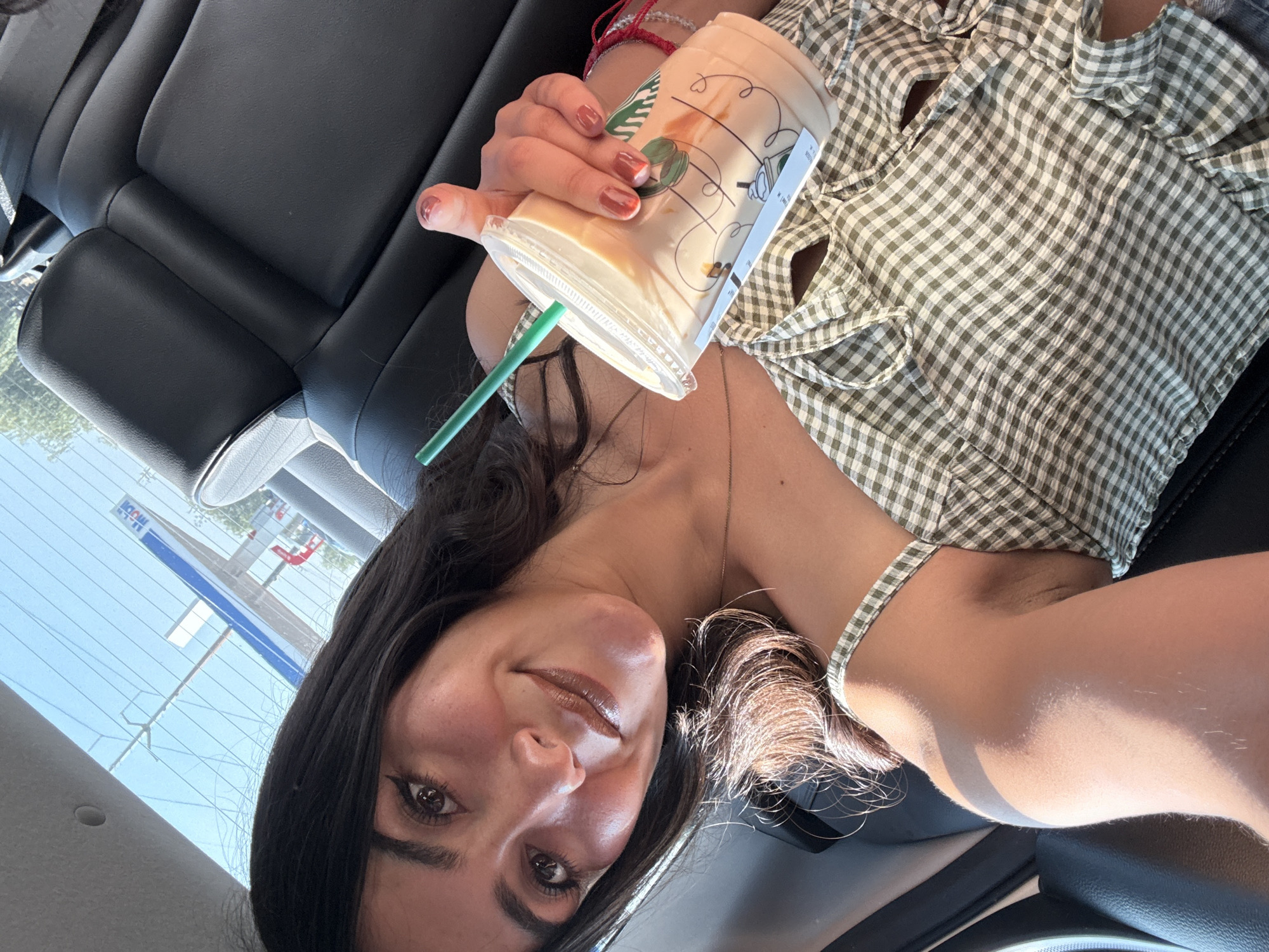 The image shows a woman in a car holding a Starbucks drink. She is wearing a green and white checkered top. The car's interior is black leather. Through the car window, a gas station sign is visible, which may be located in Santa Ana Xalmimilulco, Mexico.