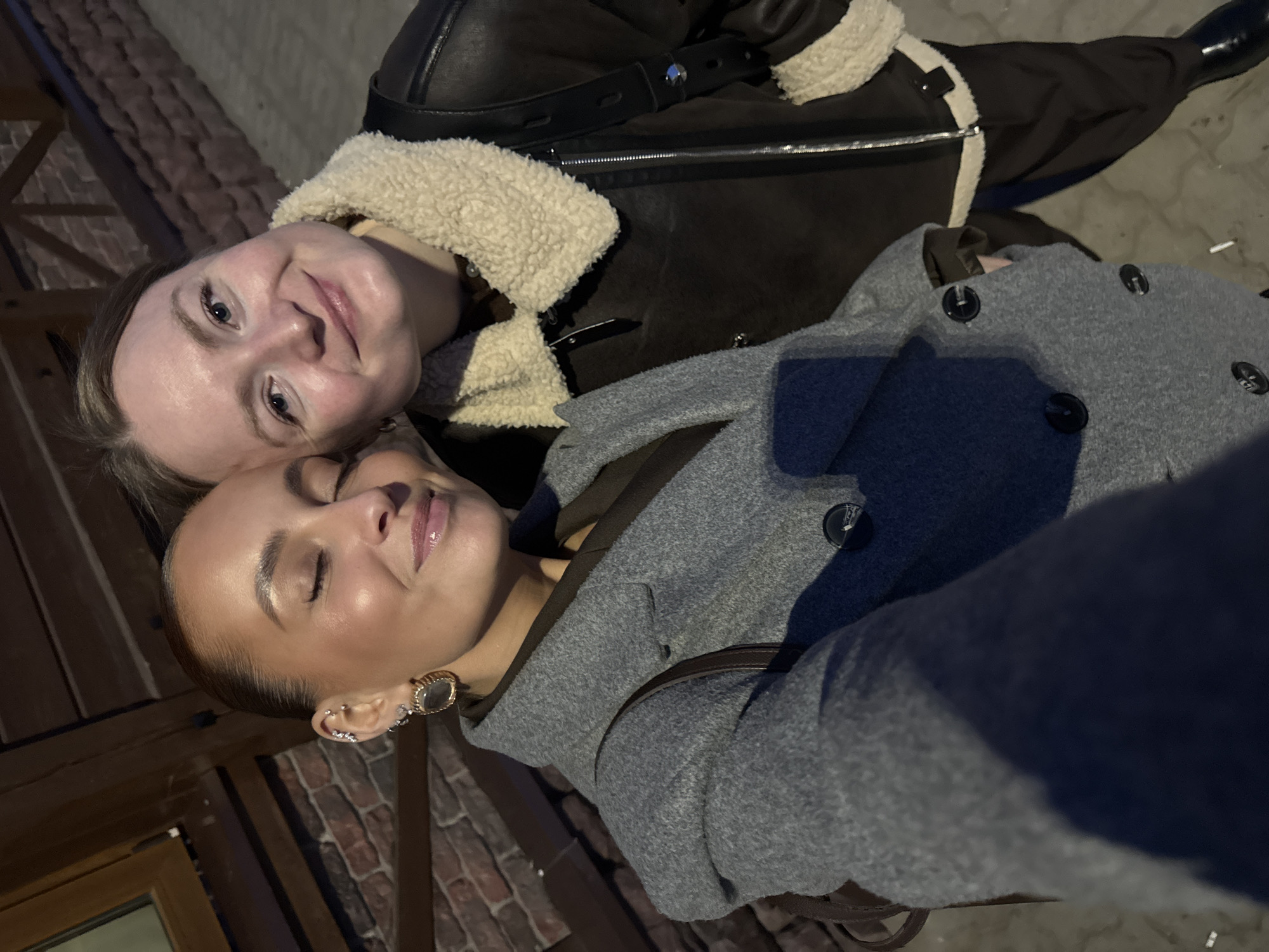 Two adult females are depicted in a close-up outdoor photograph, likely a selfie, at an indeterminate location within Chaikovsky, Russia. The woman on the left has her eyes closed, exhibiting a slight smile, and wears a grey double-breasted coat over a dark garment. Her left ear displays multiple piercings, including a large gold-toned circular earring with a reflective surface. The woman on the right has her eyes open, looking directly at the camera with a subtle smile, and wears a dark brown or black aviator-style jacket with a cream-colored sherpa collar and lining. A dark strap from a bag is visible over the left woman's right shoulder. The background features a reddish-brown brick wall and dark wooden architectural elements, suggesting a building exterior. The ground is indistinct but appears paved. The scene is illuminated by artificial light, indicative of evening or nighttime. The precise location within Chaikovsky, Russia, cannot be identified from the visual evidence.