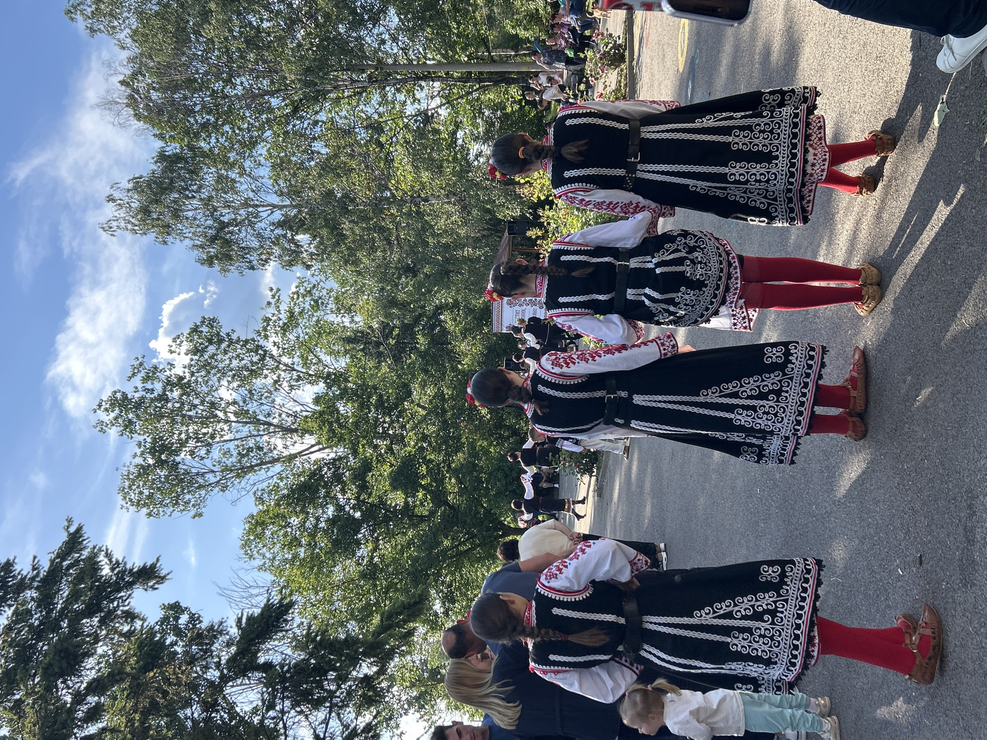 The image shows a group of people in traditional Bulgarian folk costumes in the city of Mirovyane, Bulgaria. The people in the costumes are lined up and standing on a paved road. They appear to be waiting to perform or participate in an event. Some onlookers are present on the side. The background features trees and a blue sky with clouds.