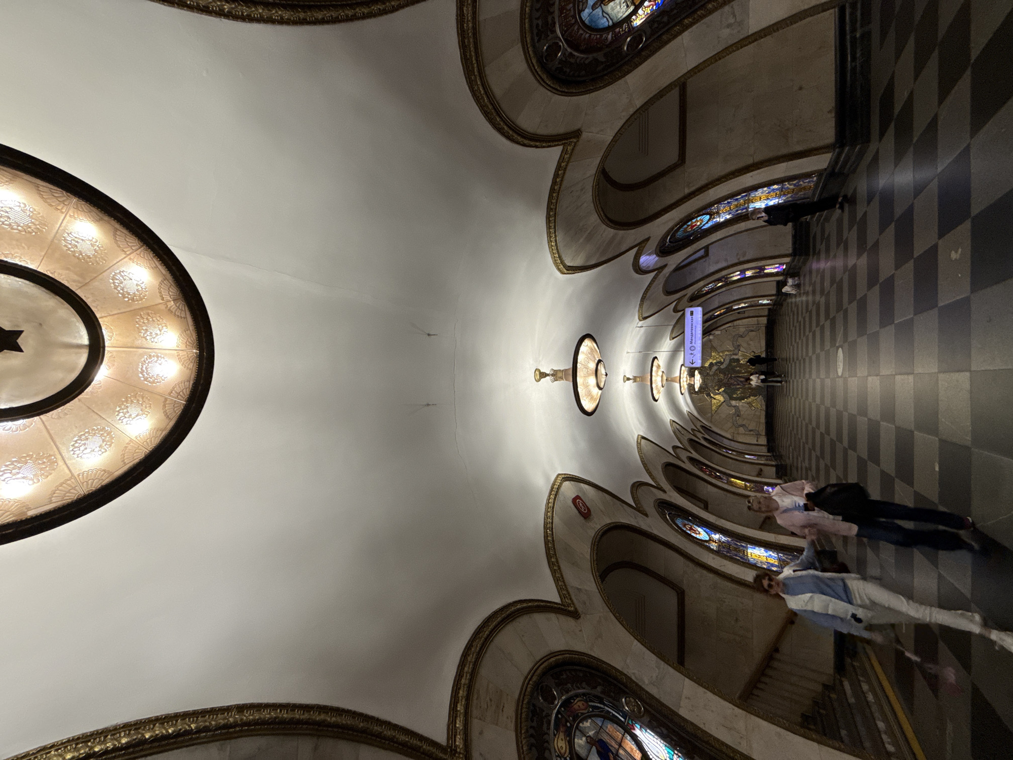 The image depicts the interior of a richly decorated Moscow Metro station. The vaulted ceiling is adorned with ornate lighting fixtures and gold trim. Along the curved walls, arched alcoves feature stained-glass windows depicting various scenes, possibly historical or allegorical. The floor is a checkered pattern of dark and light grey tiles.
In the foreground, two individuals are walking away from the camera down a corridor. The person on the left is wearing a light blue sweater, a white vest, and white pants, and is carrying a red umbrella. The person on the right is wearing a pink jacket over a white top and dark pants, and is carrying a black bag. Further down the corridor, several other individuals are visible, some appearing to stand and others walking.
A directional sign is visible on the ceiling, indicating "Менделеевская" (Mendeleevskaya), which is a station on the Serpukhovsko-Timiryazevskaya Line of the Moscow Metro. The architecture suggests a style common to many of Moscow's grand metro stations, likely dating from the Soviet era. The overall impression is one of opulence and grandeur, characteristic of the Moscow Metro system.
